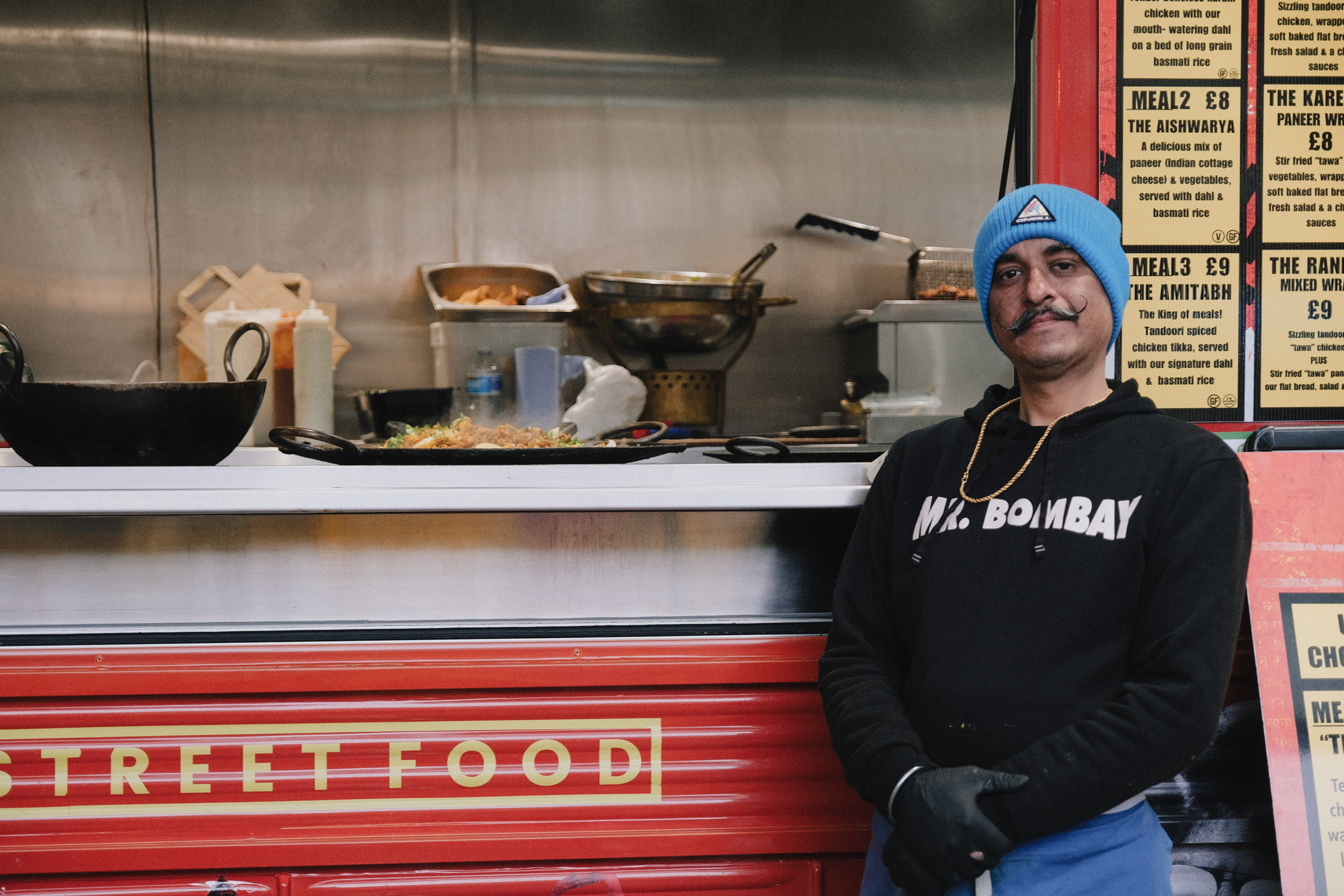 Chef in blue beanie and black "Mr. Bombay" sweatshirt stands beside red street food stall with cooking pots and yellow menu boards.