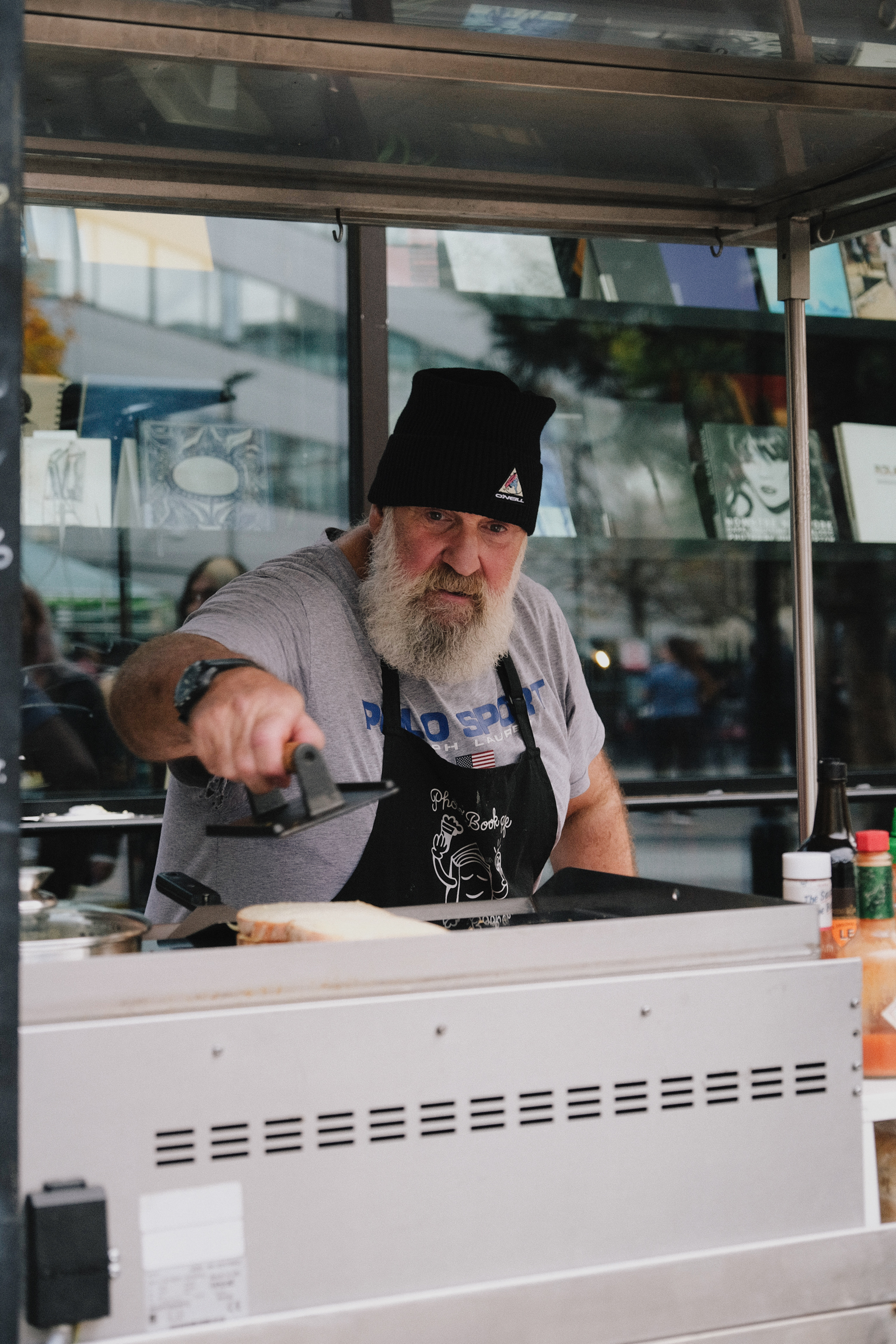 Bearded man in black beanie and apron cooking at food truck window, steam rising from grill, urban street visible behind glass.