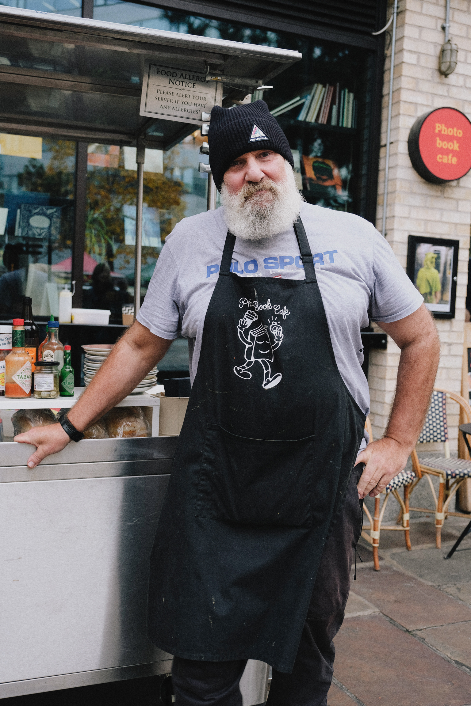 Bearded man in black apron and knit hat standing outside cafe, hands on hips, smiling at camera.