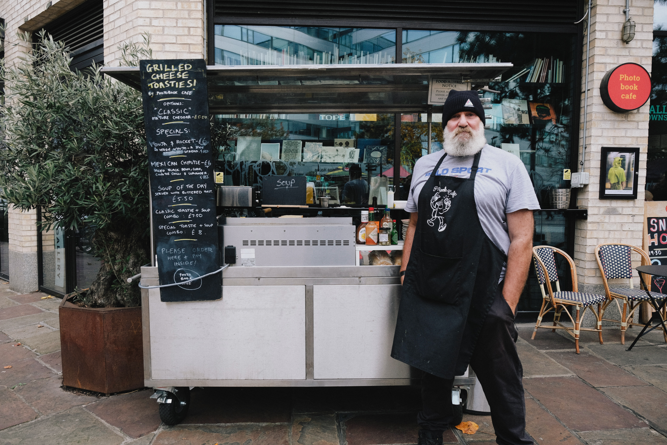 Bearded man in black apron stands beside white food cart with chalkboard menu outside café with wicker chairs and potted plant.