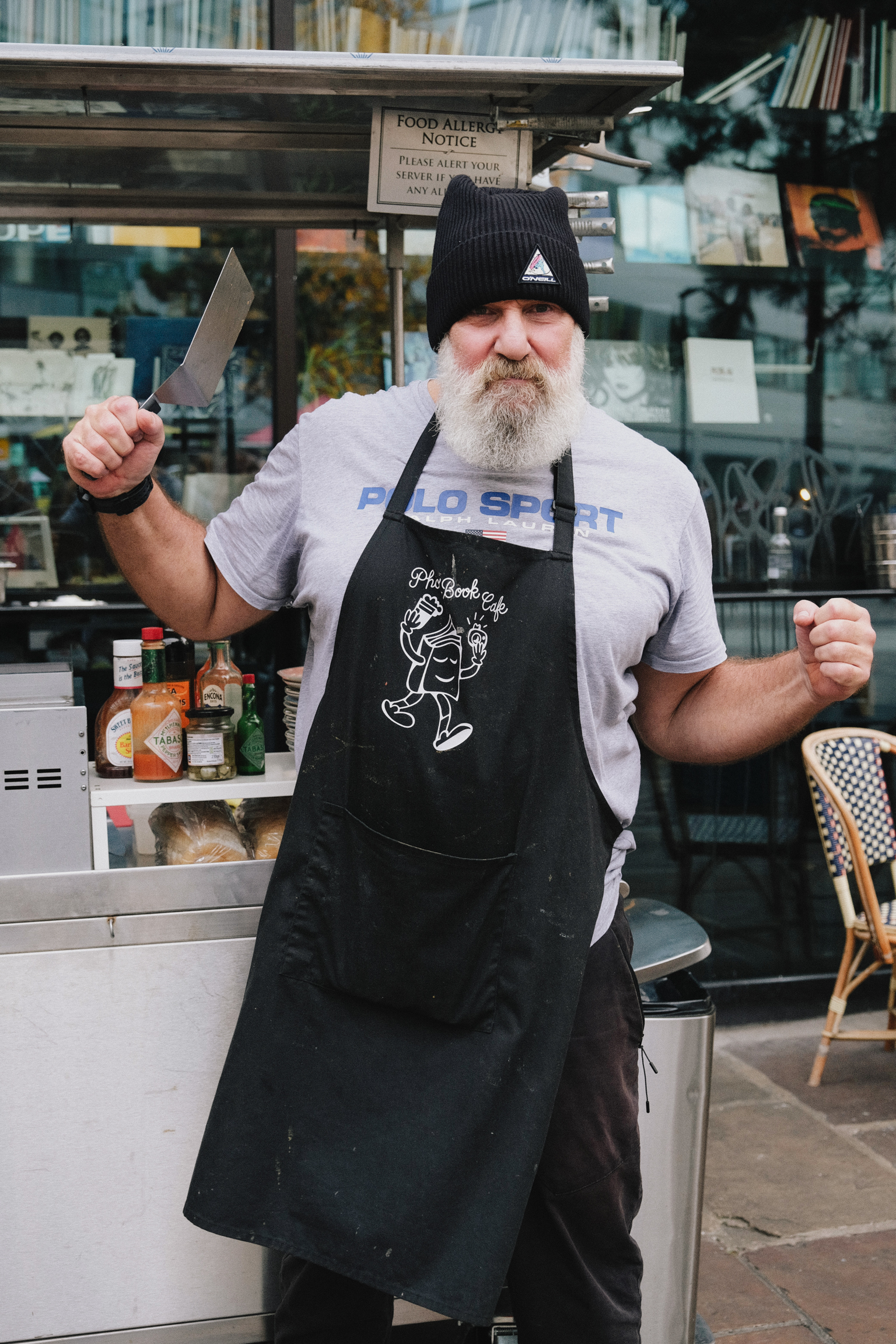 Bearded chef in black beanie and apron holds knife whilst standing outside shop with books visible in window behind him.