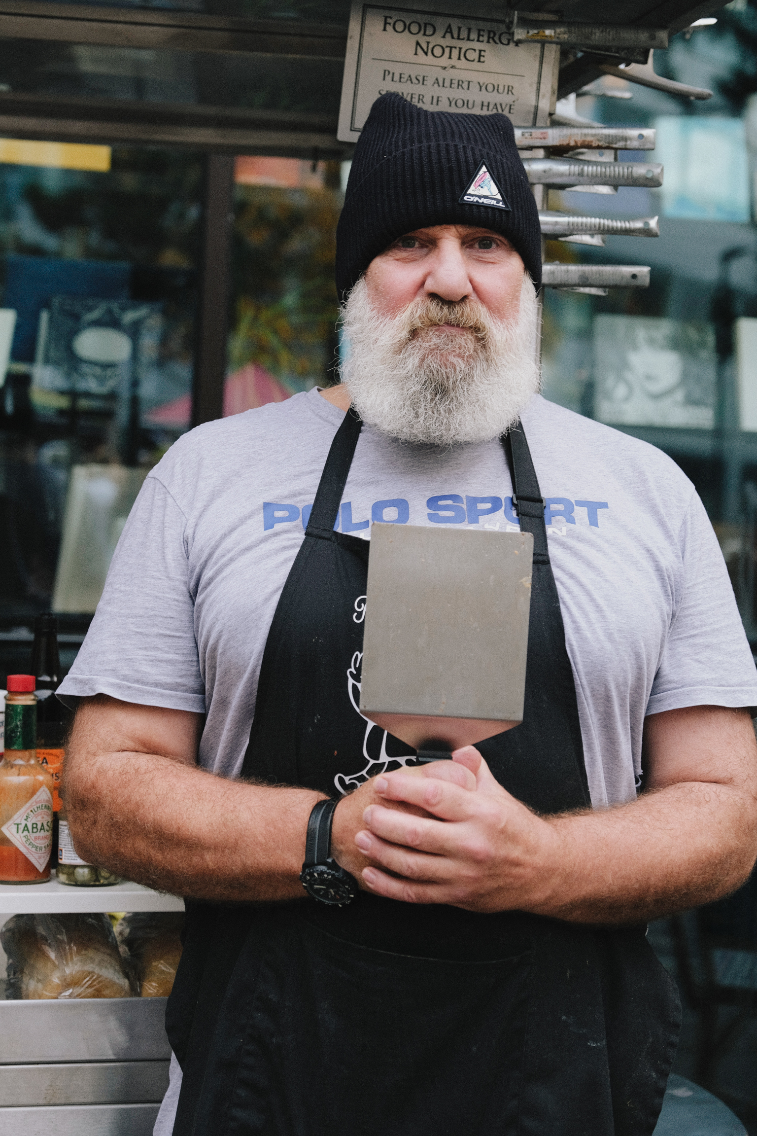 Bearded man in black beanie and apron standing outside food establishment with "Food Allergy Notice" sign visible in window behind him.