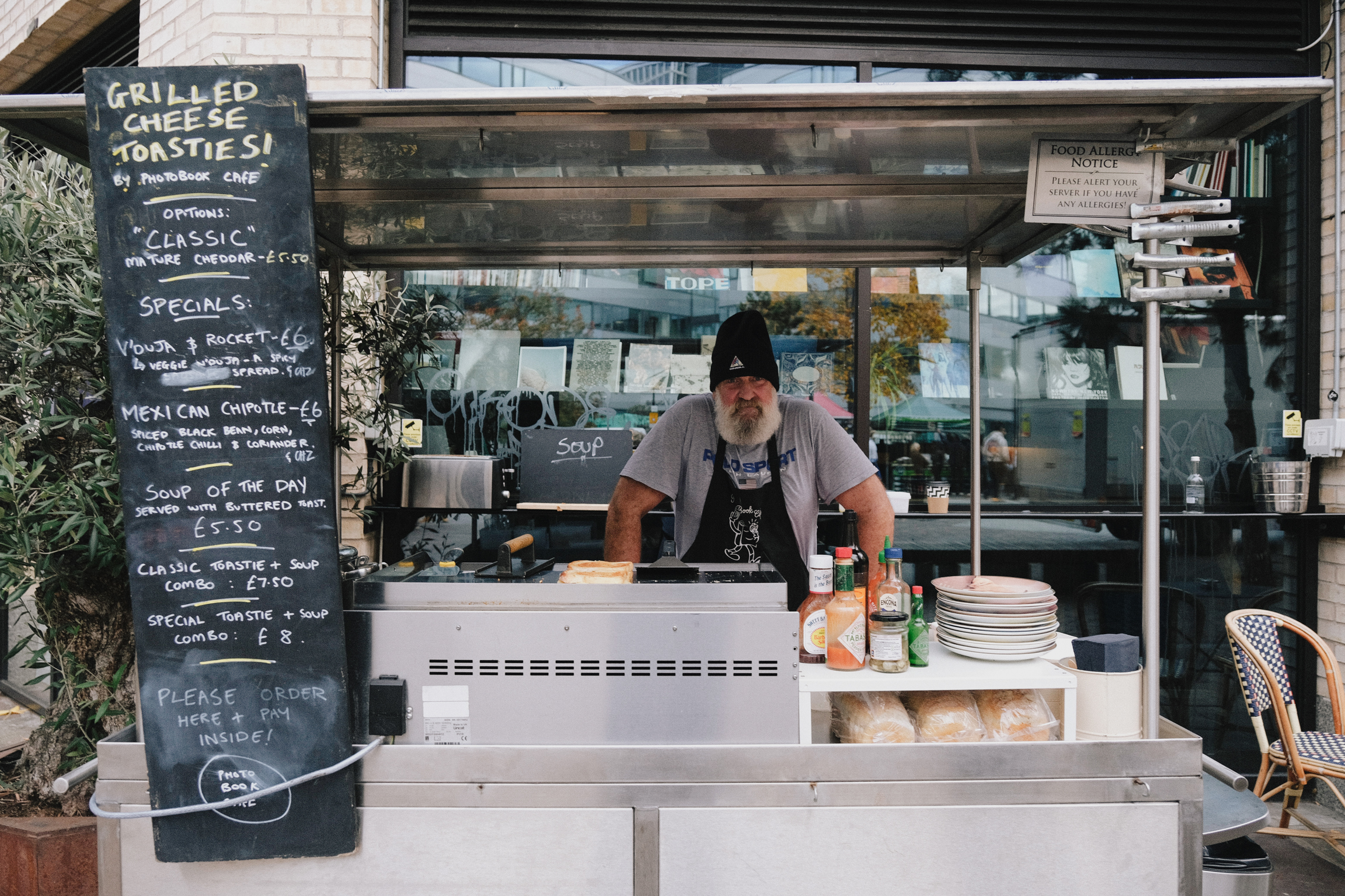 Chef in black beanie working at outdoor food stall with blackboard menu, white counter, stacked plates, and glass service window.