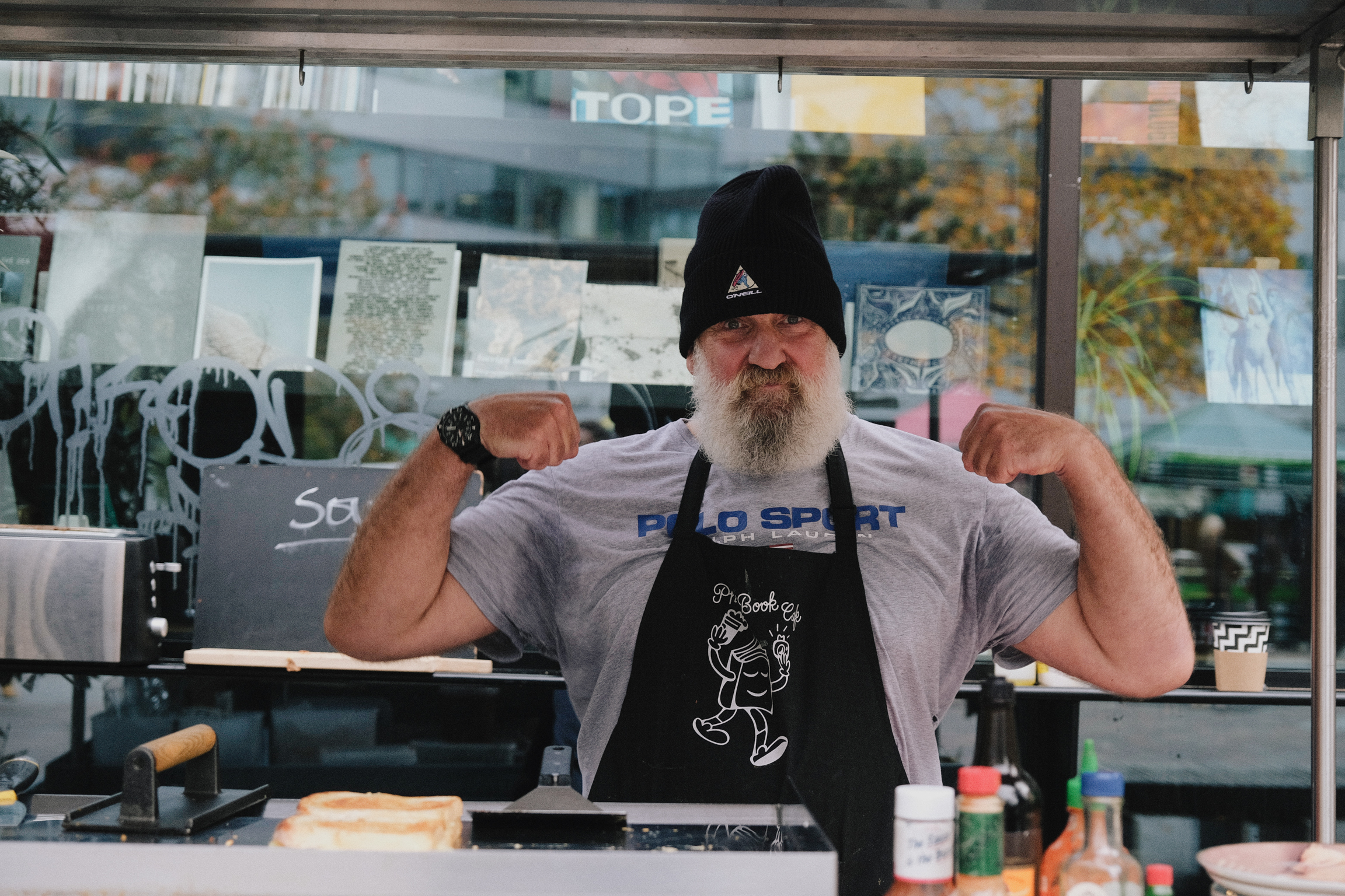 Bearded man in black beanie and apron flexing arms behind deli counter, with "HOPE" sign visible in shop window behind him.