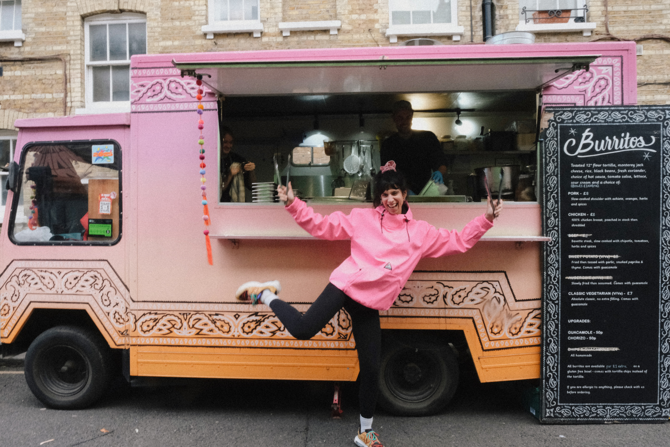 Person in pink hoodie with arms raised standing beside pink and orange food truck with decorative patterns and menu board.