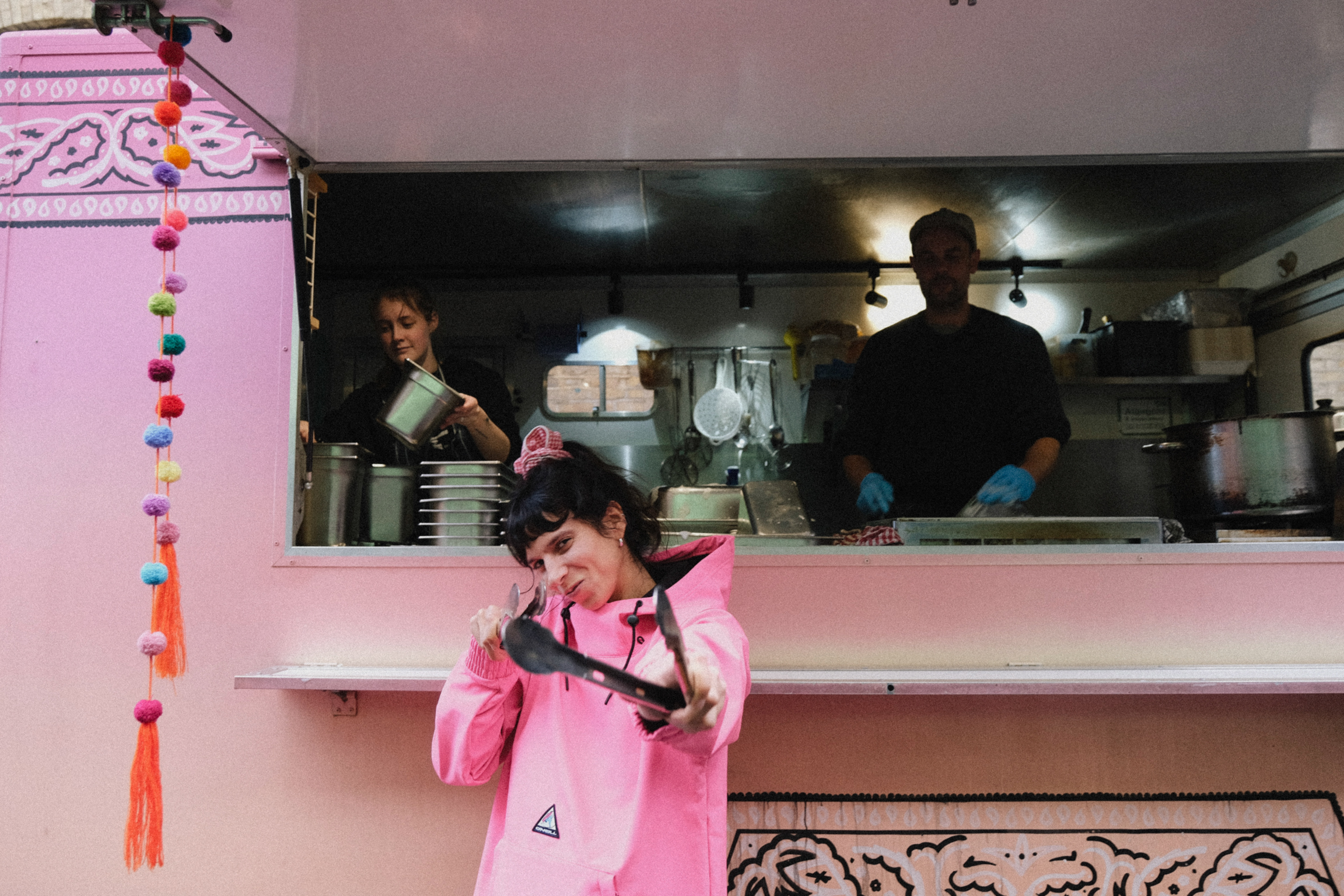 Woman in pink hoodie on phone at restaurant counter with staff working in kitchen behind service window. Colourful pom-pom decorations hang nearby.