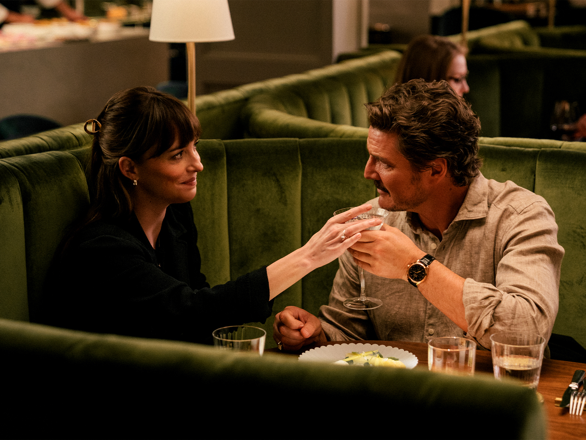 Woman in black top and man in beige shirt sitting in green booth, woman touching man's hands across table with glasses.