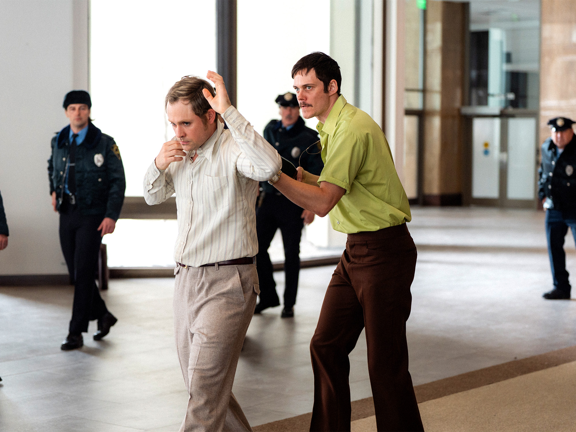 Two men in corridor, one in beige shirt covering face, other in yellow shirt behind him. Police officers in dark uniforms visible in background.