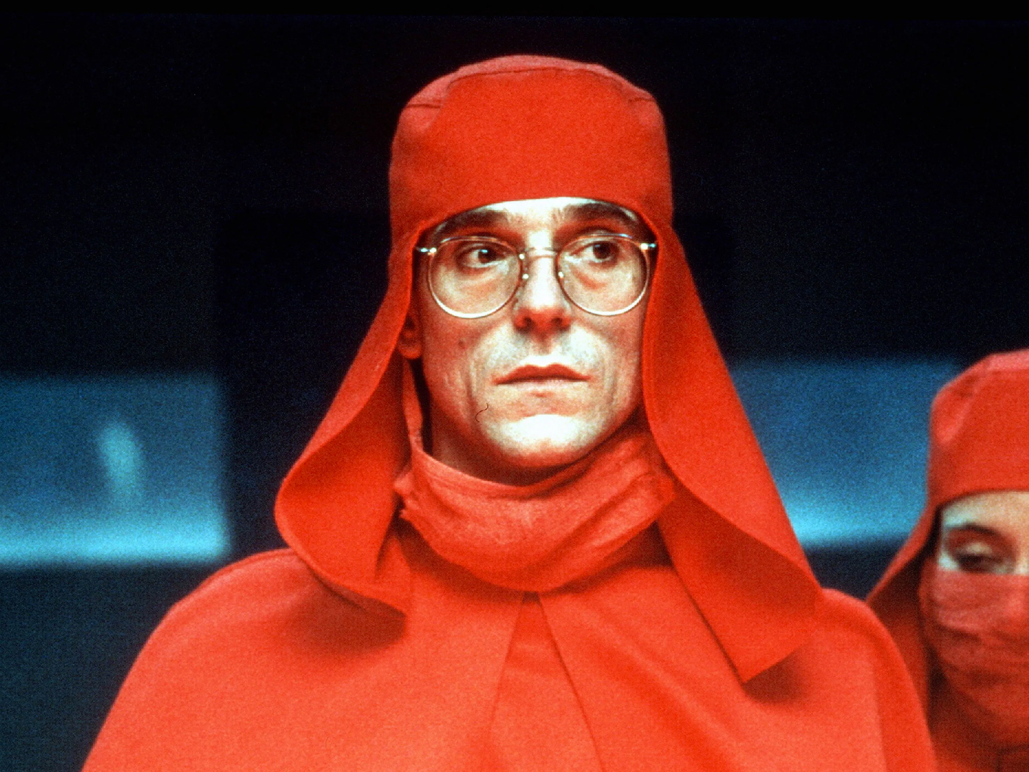 A person wearing a red hooded robe and glasses against a dark background.