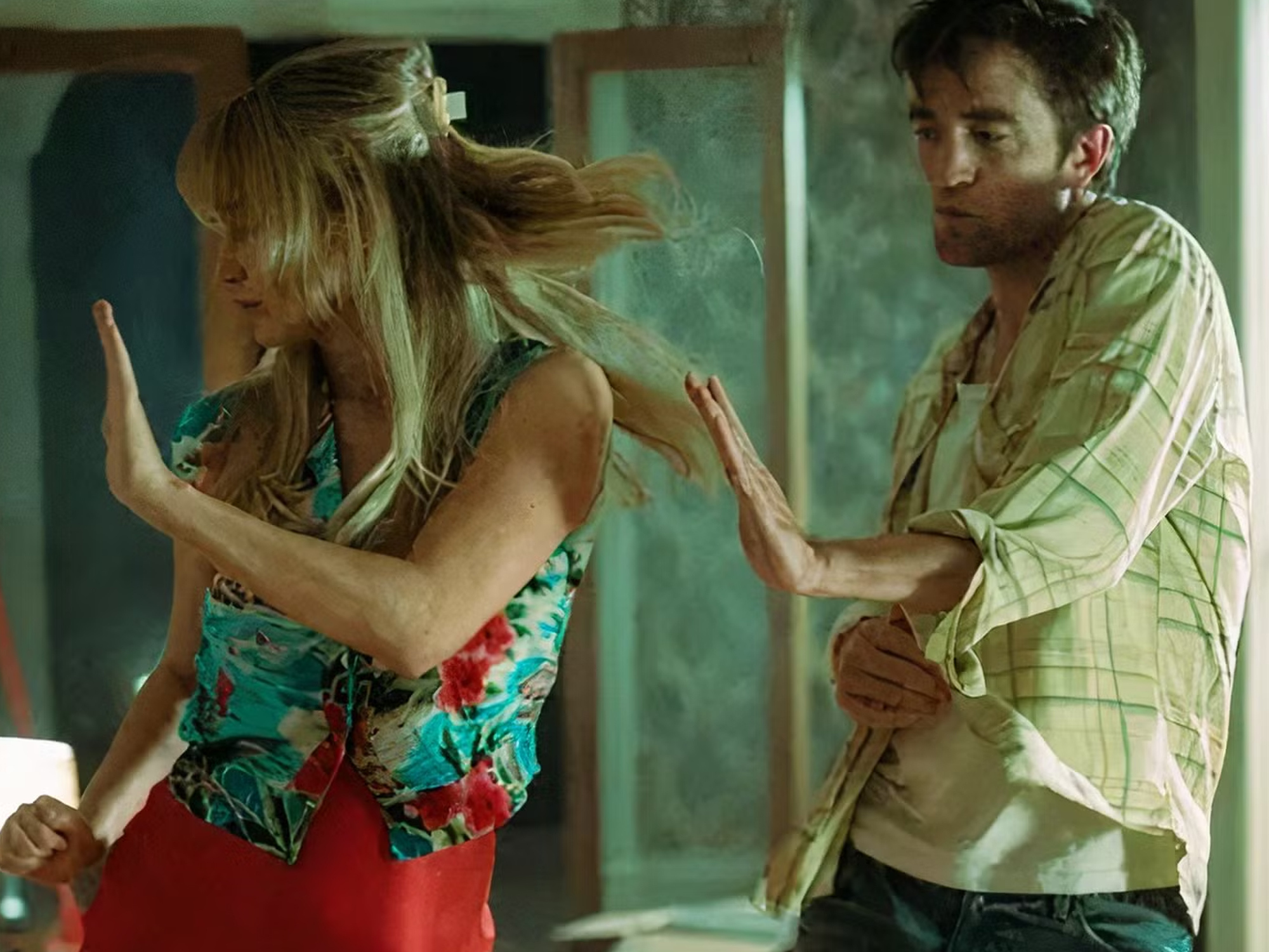 Two people, a man and a woman, locked in an intense embrace, appearing to be in distress. The woman has long blonde hair and is wearing a floral printed dress, while the man has short dark hair and is wearing a light-coloured shirt.