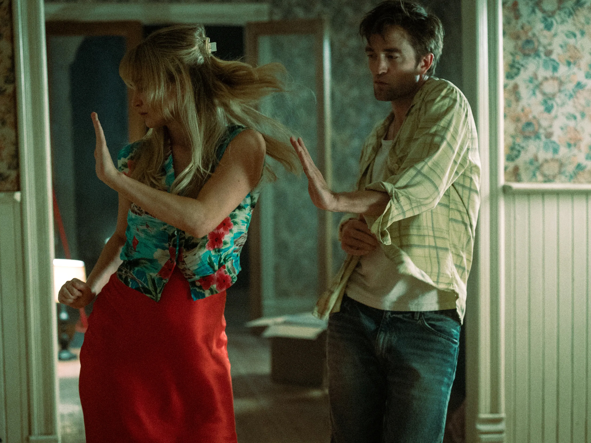 Jennifer Lawrence, a blonde woman a floral dress, and Robert Pattinson in a yellow checked shirt and jeans, dance in a room with patterned wallpaper.