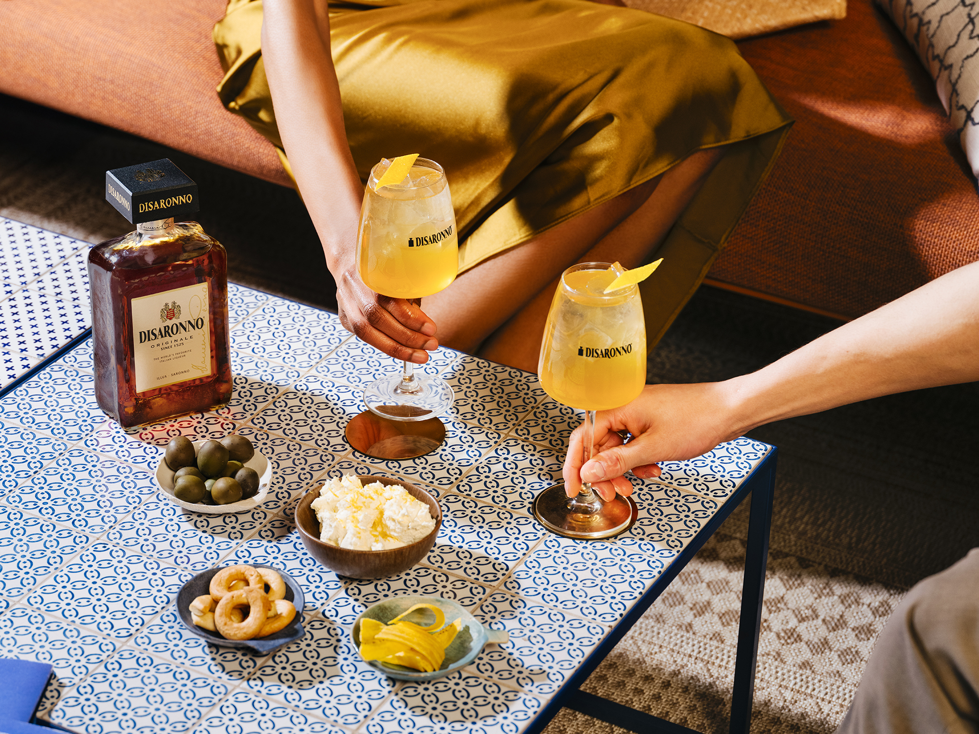 Hands holding cocktail glasses filled with Disaronno cocktails, surrounded by olives, lemon slices, and other appetizers on a blu patterned table.