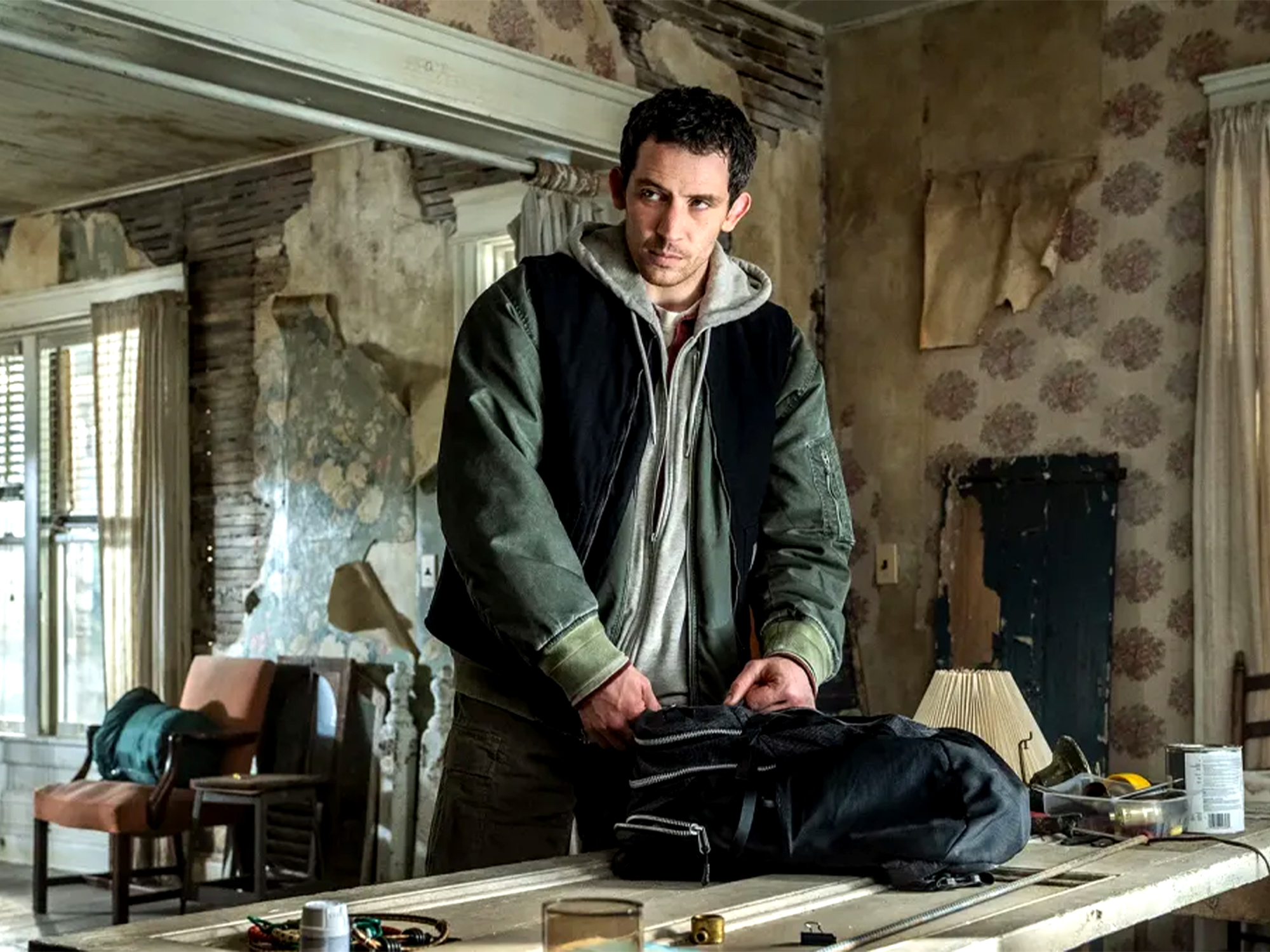 Man in green jacket and hoodie kneeling on wooden floor in rundown room with peeling walls, exposed beams, and renovation materials.