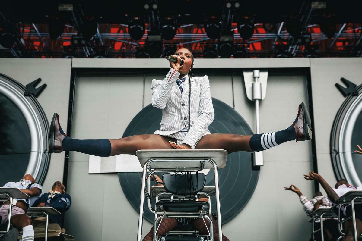 Performer in white suit doing splits above crowd on industrial set with circular green backdrop and metal framework.
