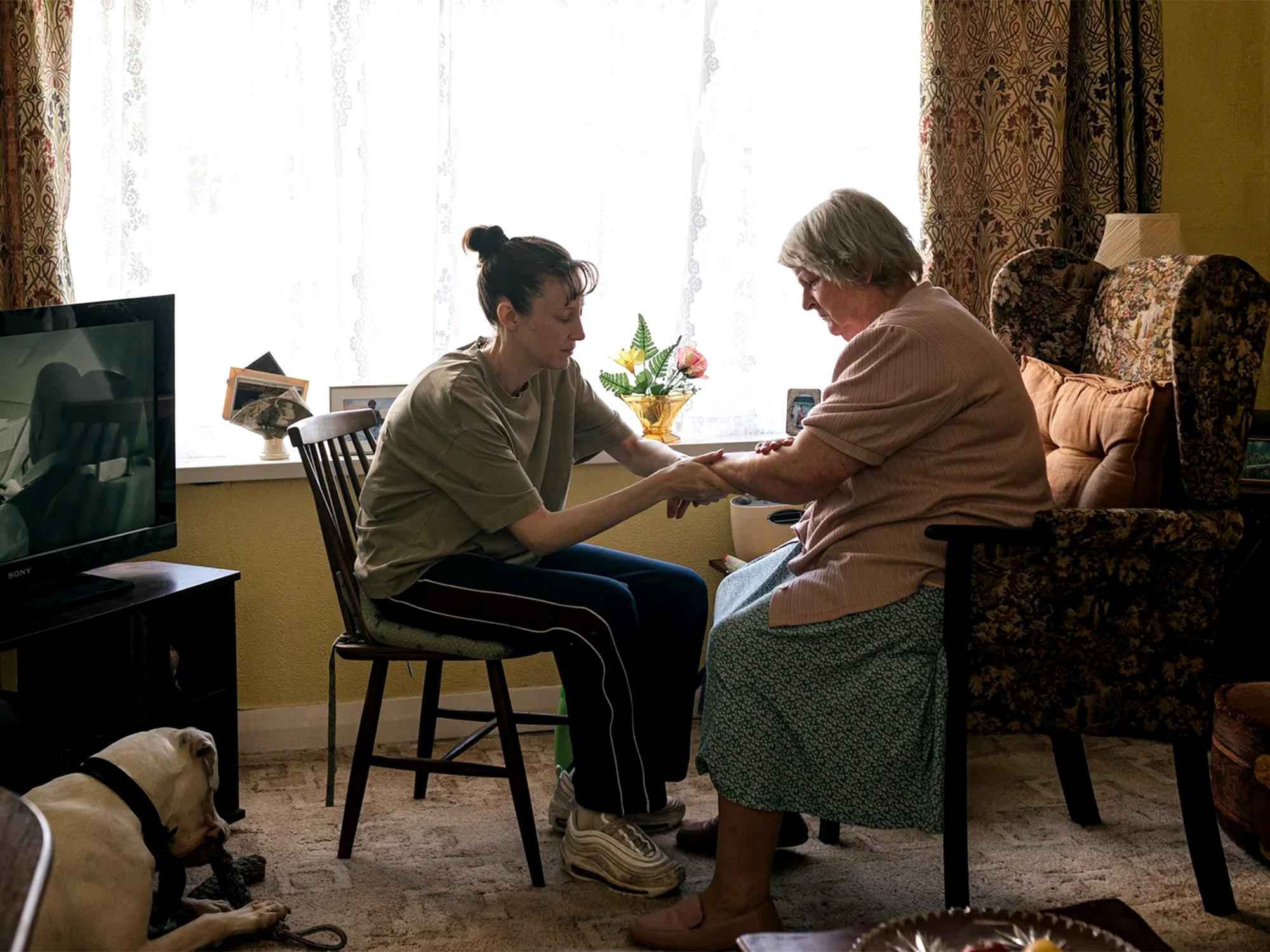 Young woman in green cardigan sits beside elderly woman on patterned sofa, holding hands in warm living room with television and dog.