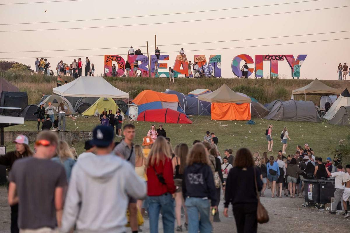 Large colourful "DREAM CITY" letters on hillside above camping area with multicoloured tents and crowds of people walking below.