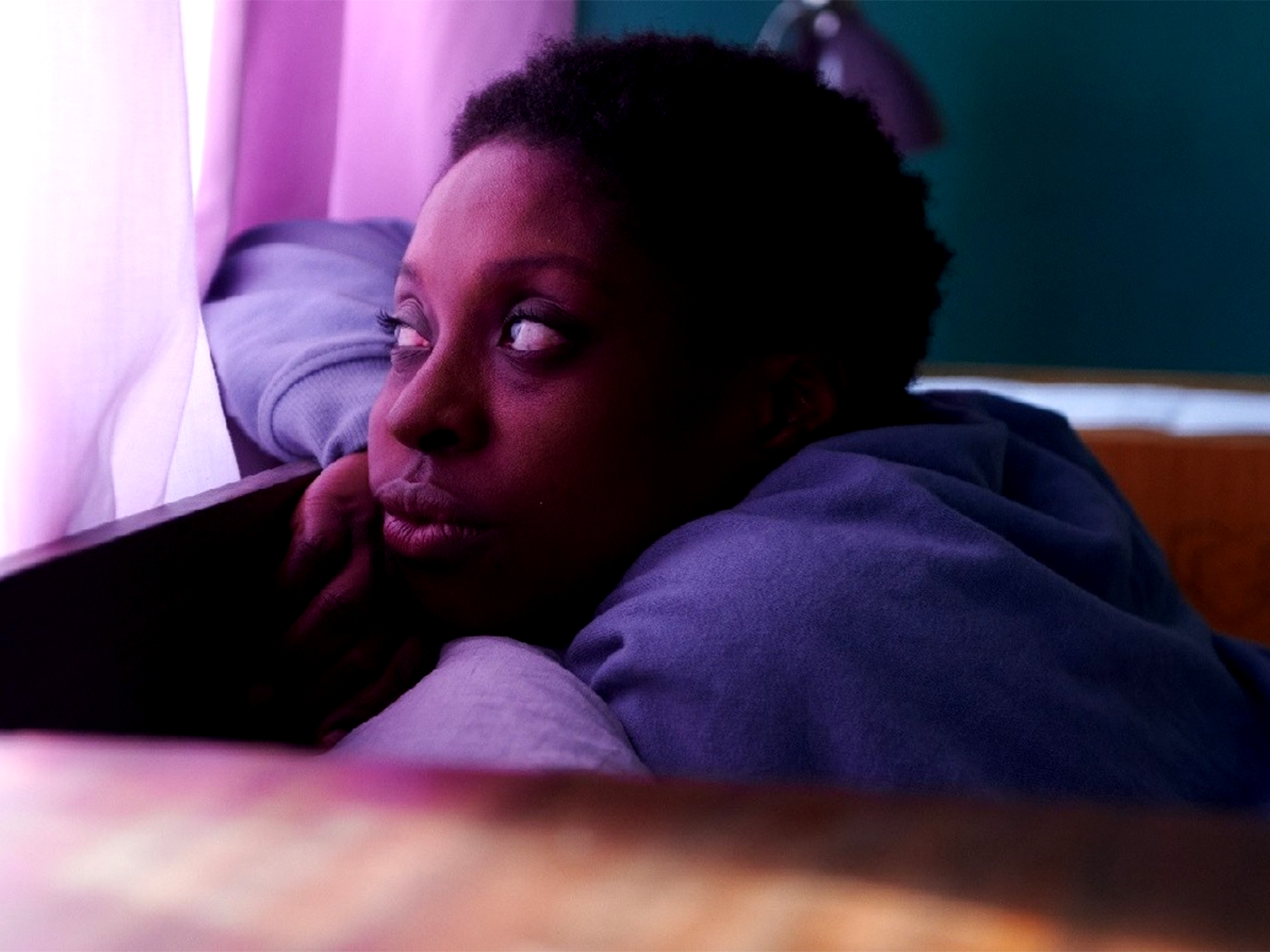 Person with dark curly hair wearing purple hoodie, lying down with hand near face, illuminated by pink and blue lighting.