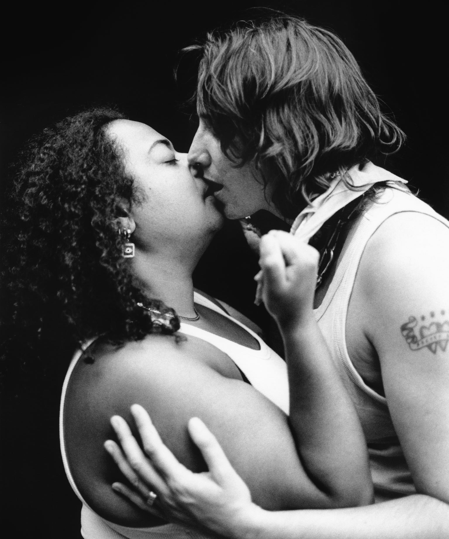 Black and white image of two people kissing intimately, one with curly hair wearing earrings, the other with straight hair.