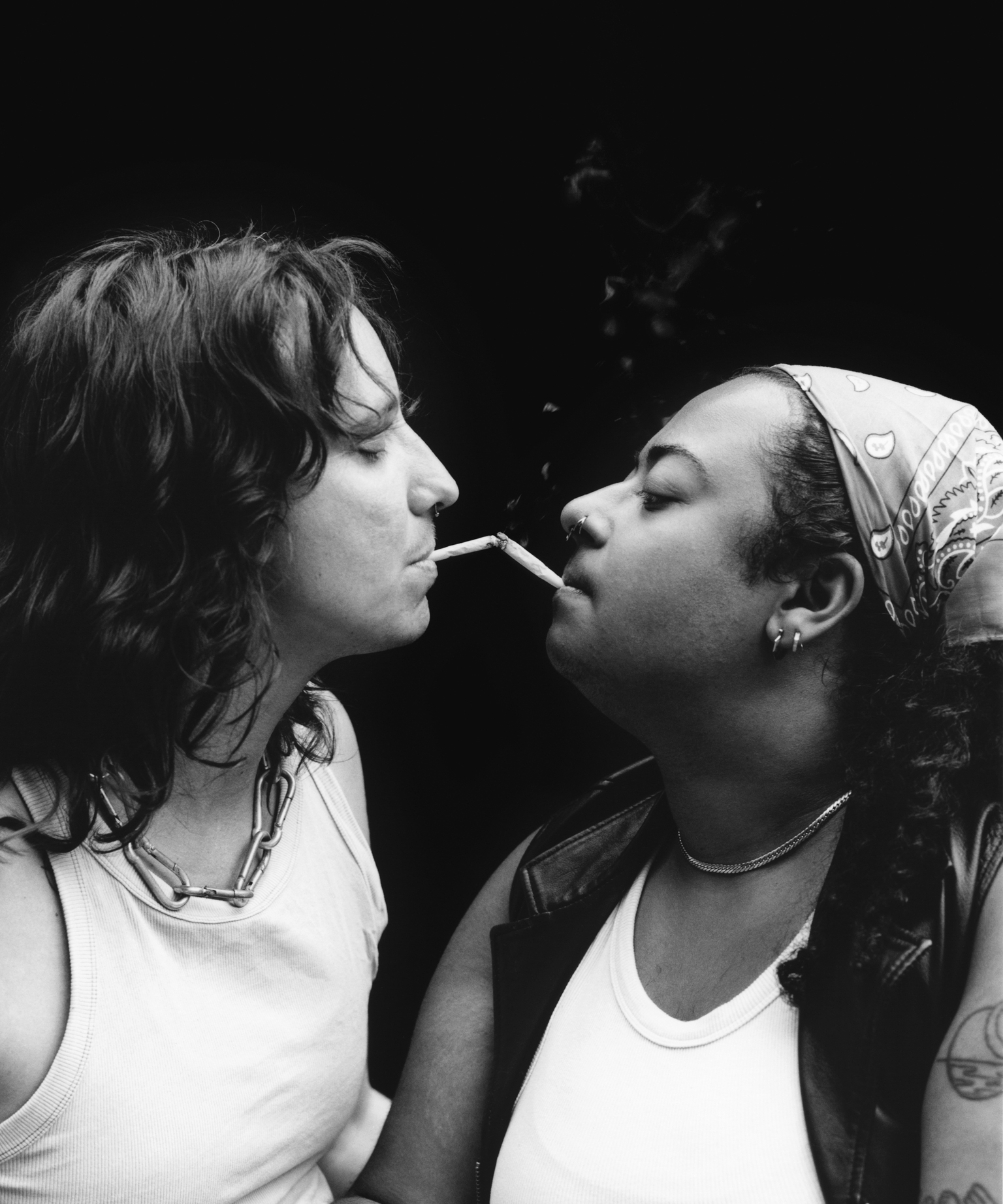 Black and white image of two people sharing a cigarette, one with curly hair in white vest, other wearing headscarf, close together.
