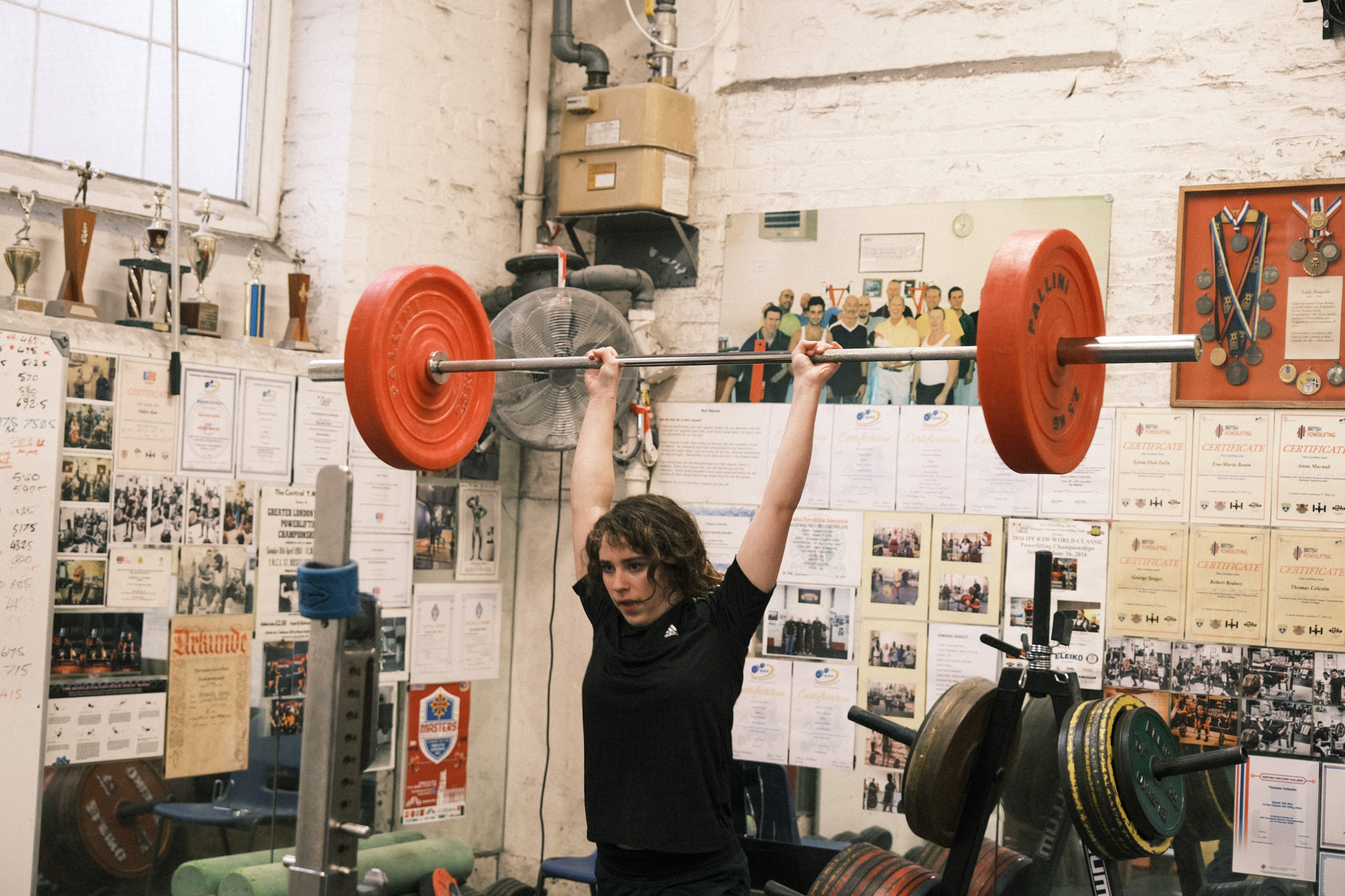 Person with curly hair lifting barbell with red weights in cluttered garage gym with newspaper clippings on walls.