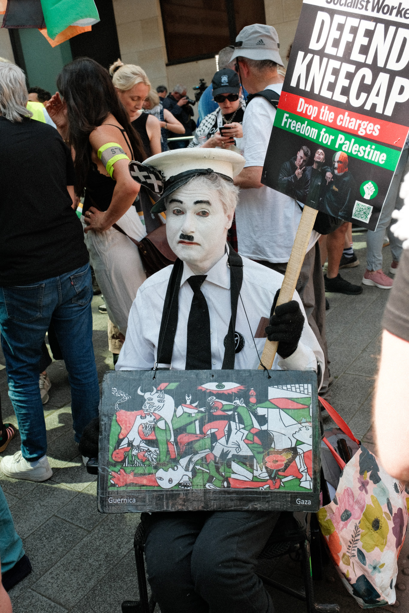 Protestor in wheelchair holding placard with Guernica painting, "Defend Kneecap, Drop the charges, Freedom for Palestine" banner