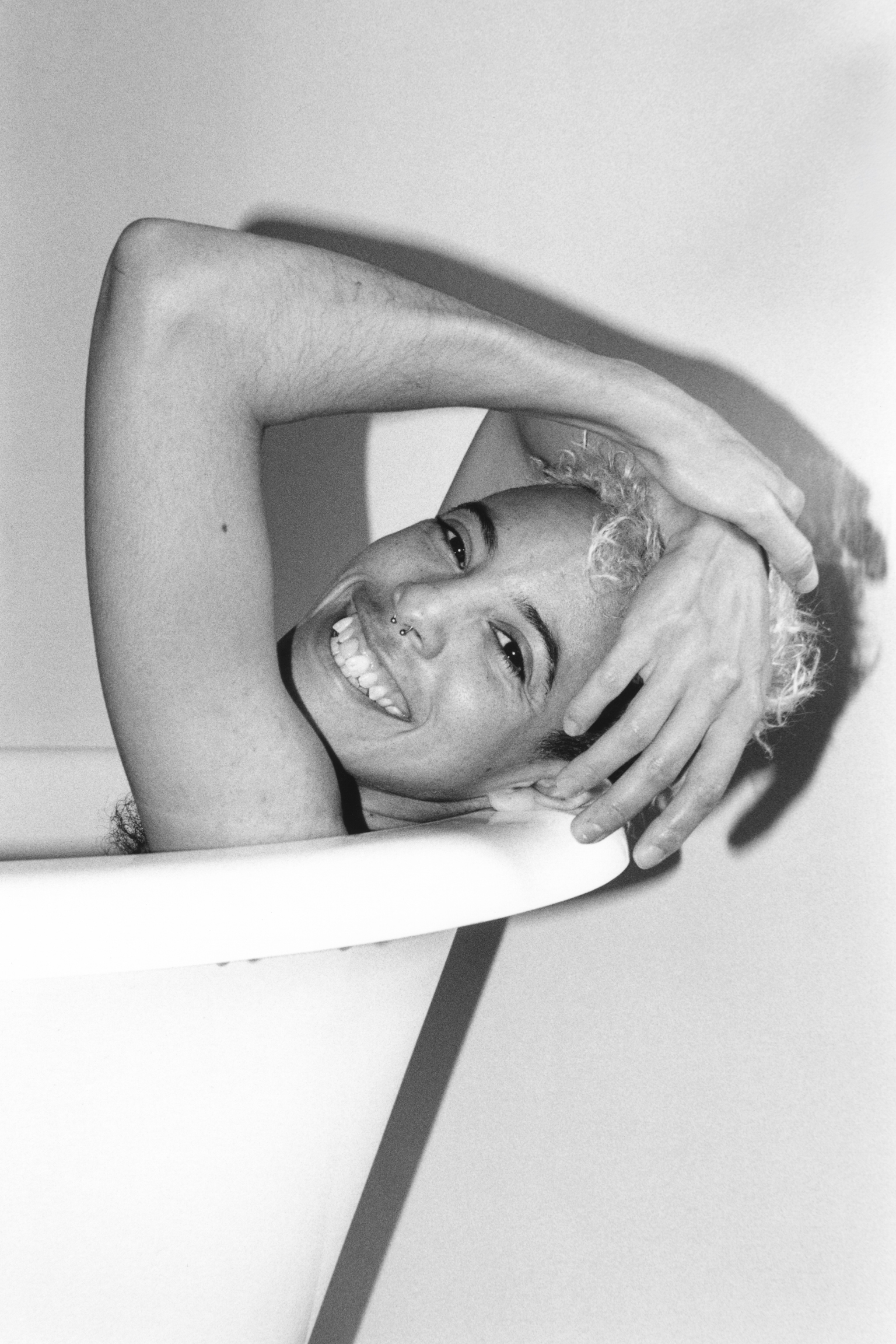 Black and white portrait of smiling woman with arms raised above head, hands in hair, leaning against white surface.