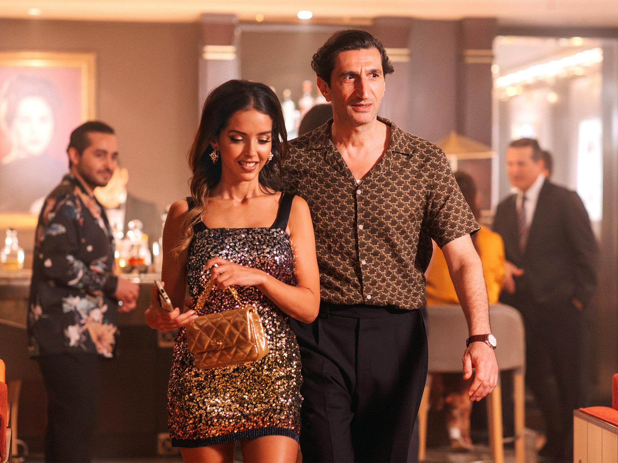 A woman in a gold sequinned dress and a man in a patterned shirt stand together in a room with other people in the background.