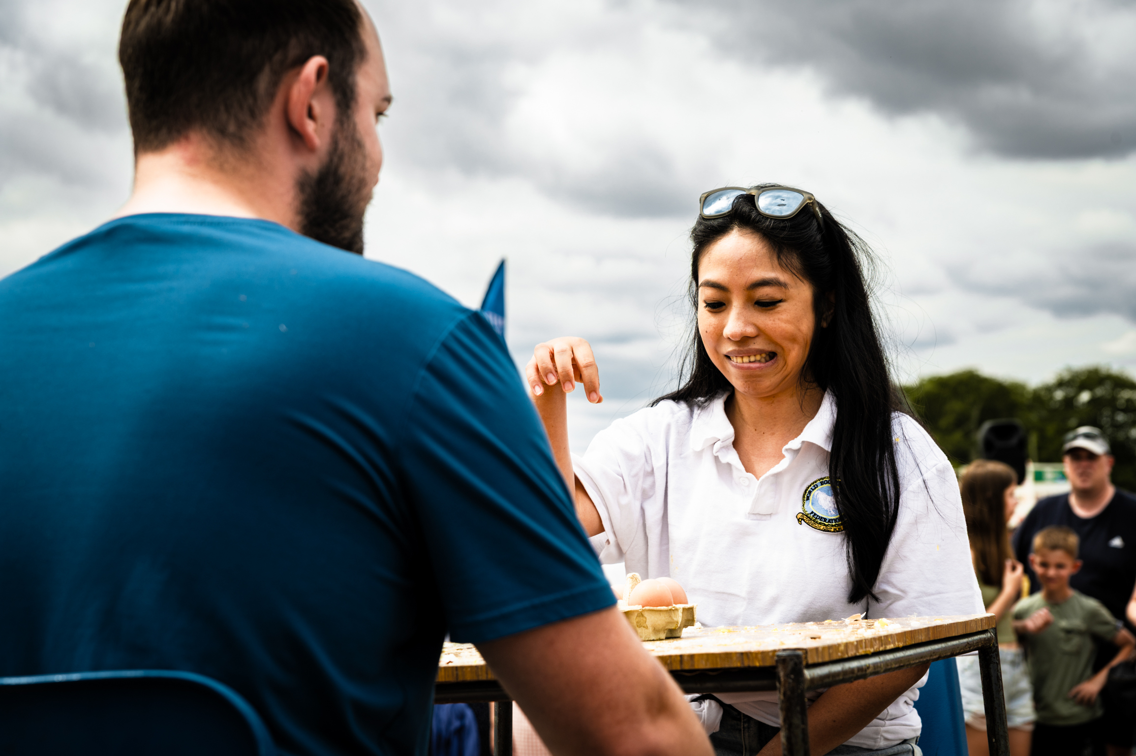 Woman in white shirt with sunglasses on head speaks to man in blue shirt at outdoor table with crowd in background.