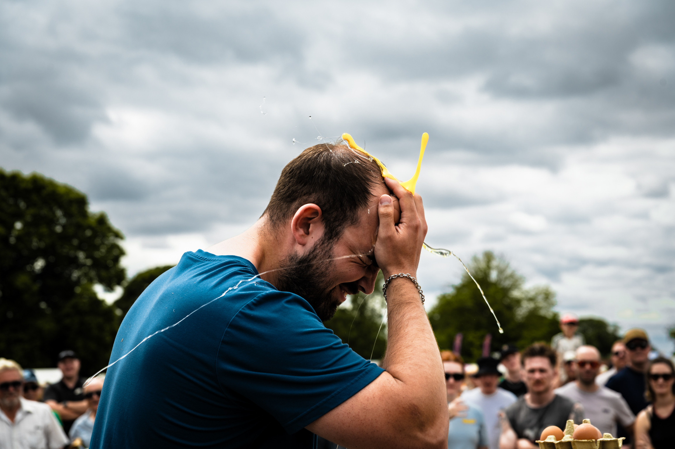 Man in blue shirt holding yellow pencil to his head, crowd and trees in background under cloudy sky.