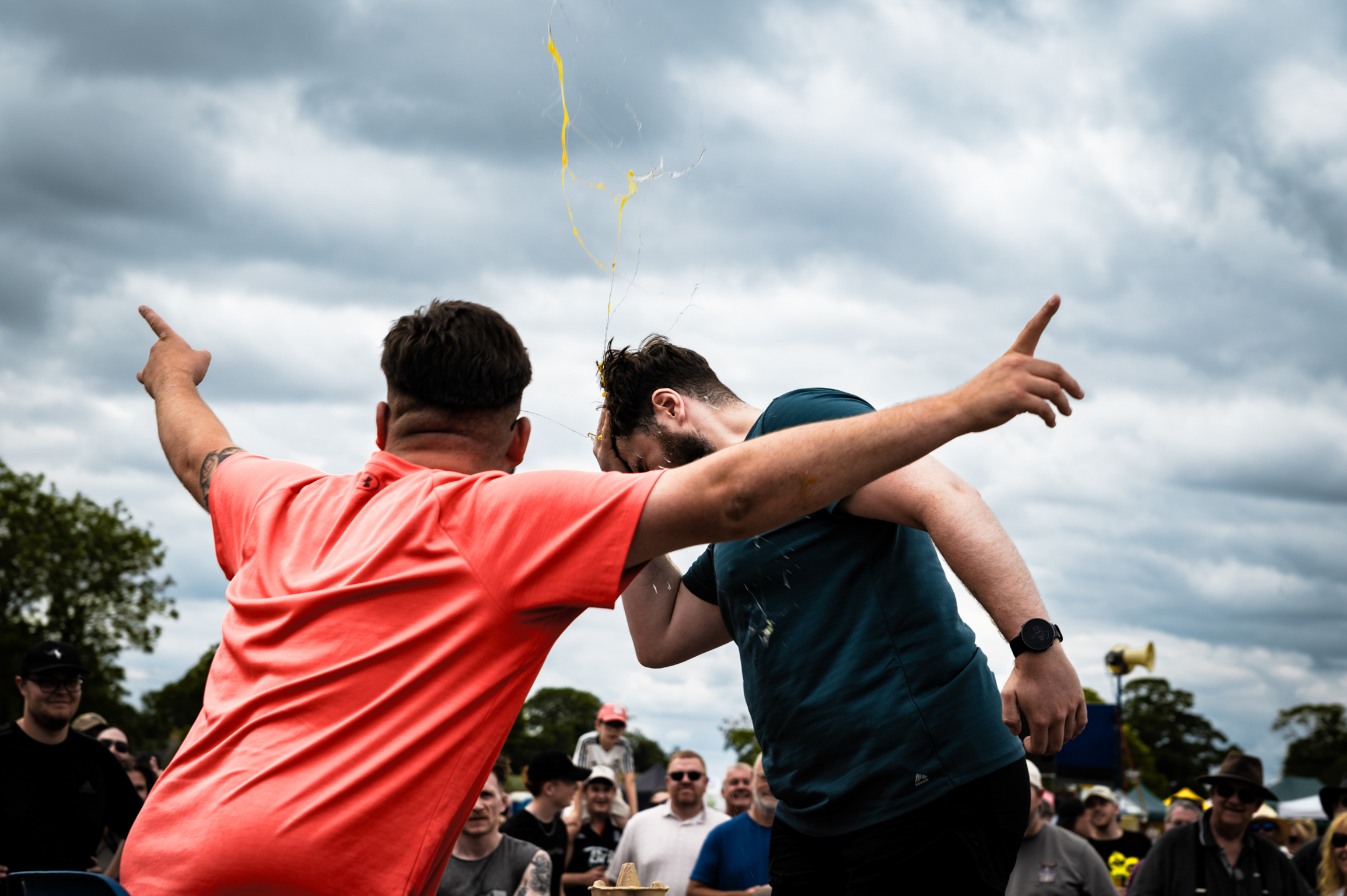 Two men with arms around each other pointing in opposite directions, one wearing orange shirt, other in dark blue, crowd behind them.