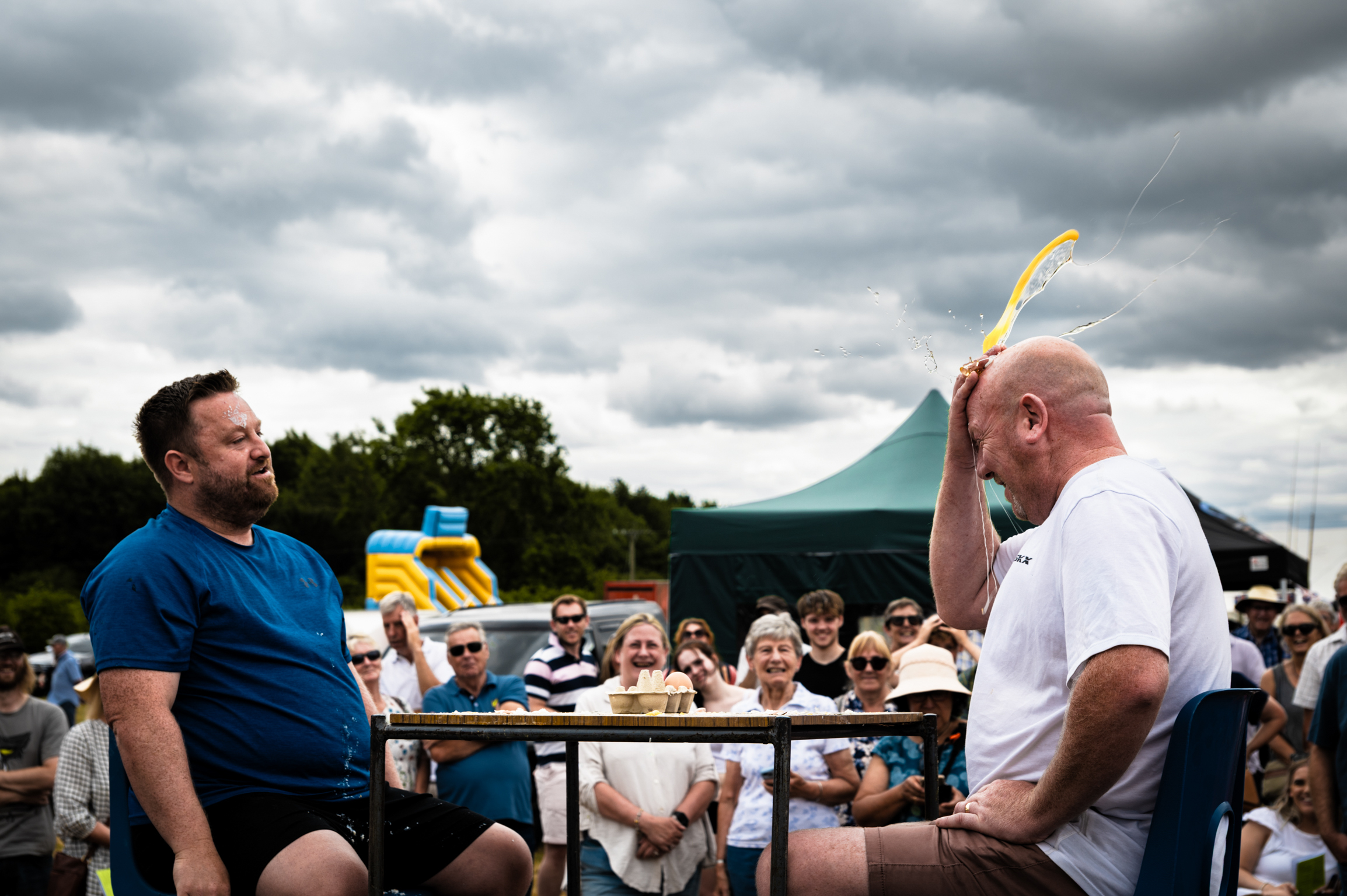 Two men at outdoor event, one in blue shirt seated, another in white holding yellow object. Crowd watching behind barriers under cloudy sky.