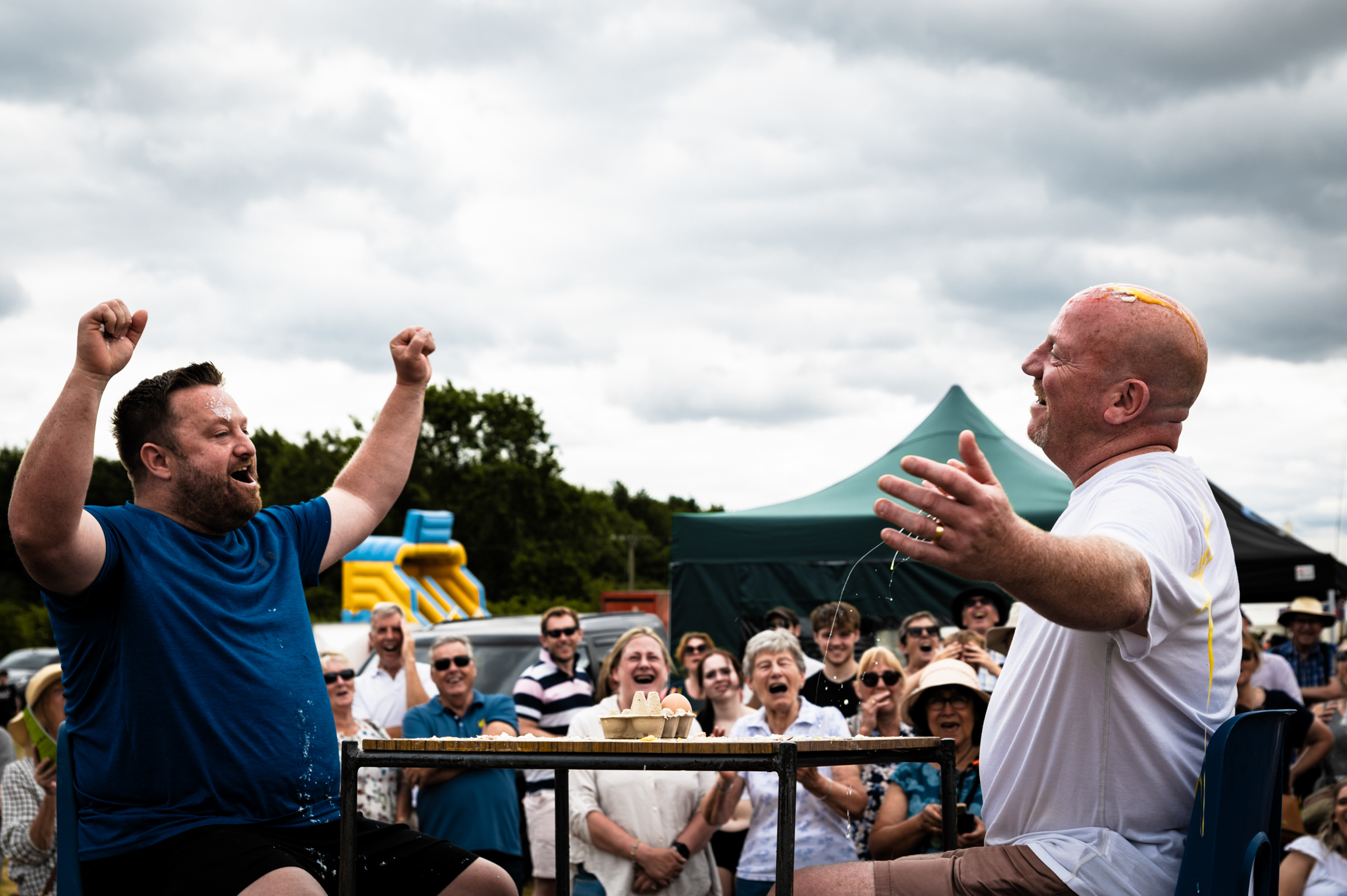 Two men at outdoor event, one in blue shirt celebrating with raised fists, bald man in white holding green object, crowd watching behind barrier.