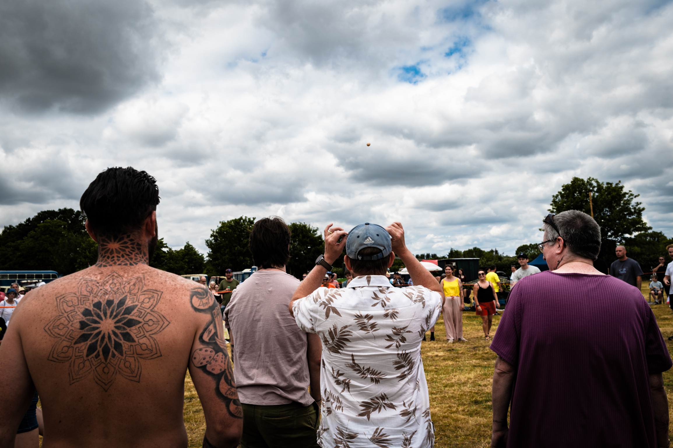 Crowd at outdoor event under cloudy sky, man with geometric back tattoo in foreground, people wearing casual shirts and caps.