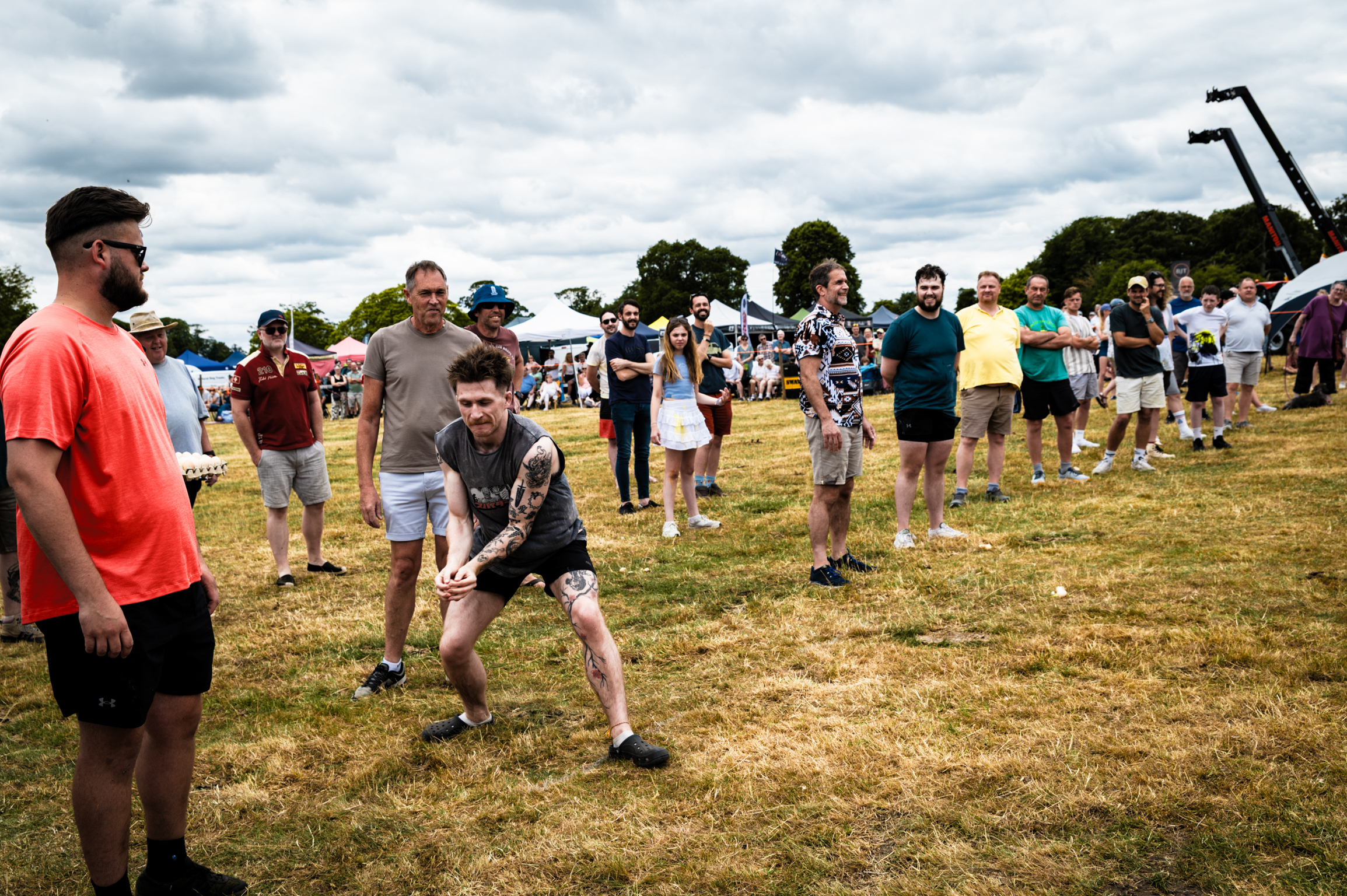 Man in grey shirt crouching in throwing position on grass field whilst crowd watches at outdoor event under cloudy sky.