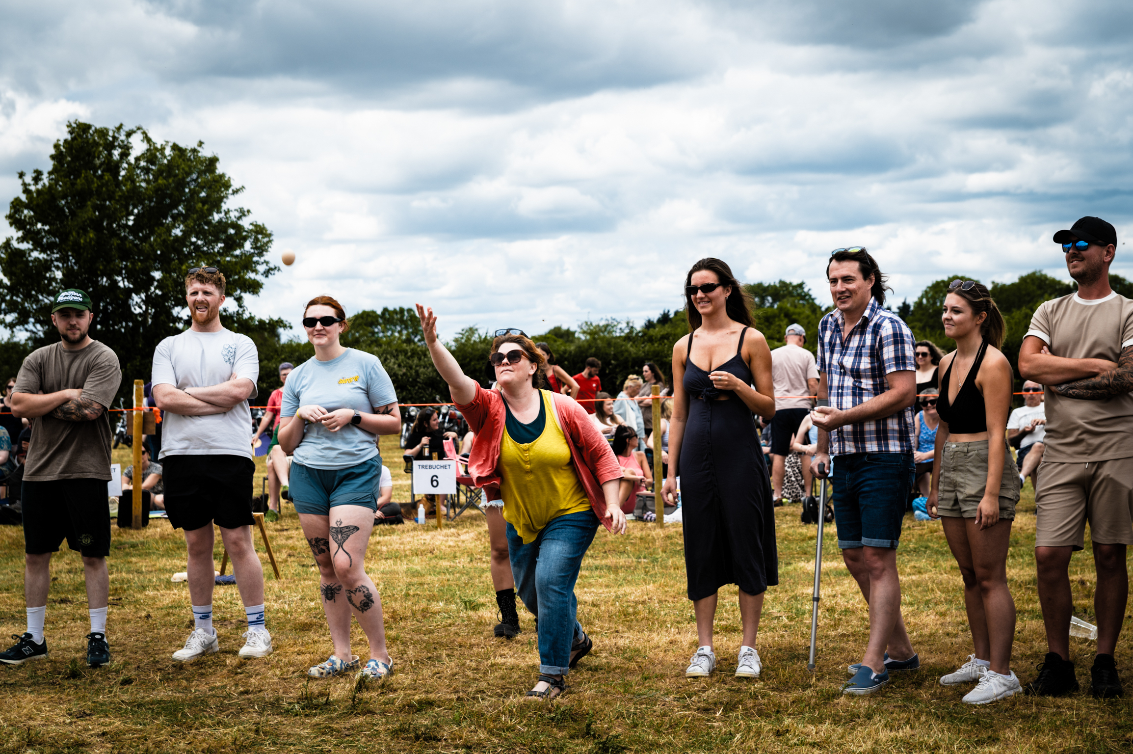 Group of people standing in line on grass field, woman in yellow top waving, cloudy sky and trees in background.