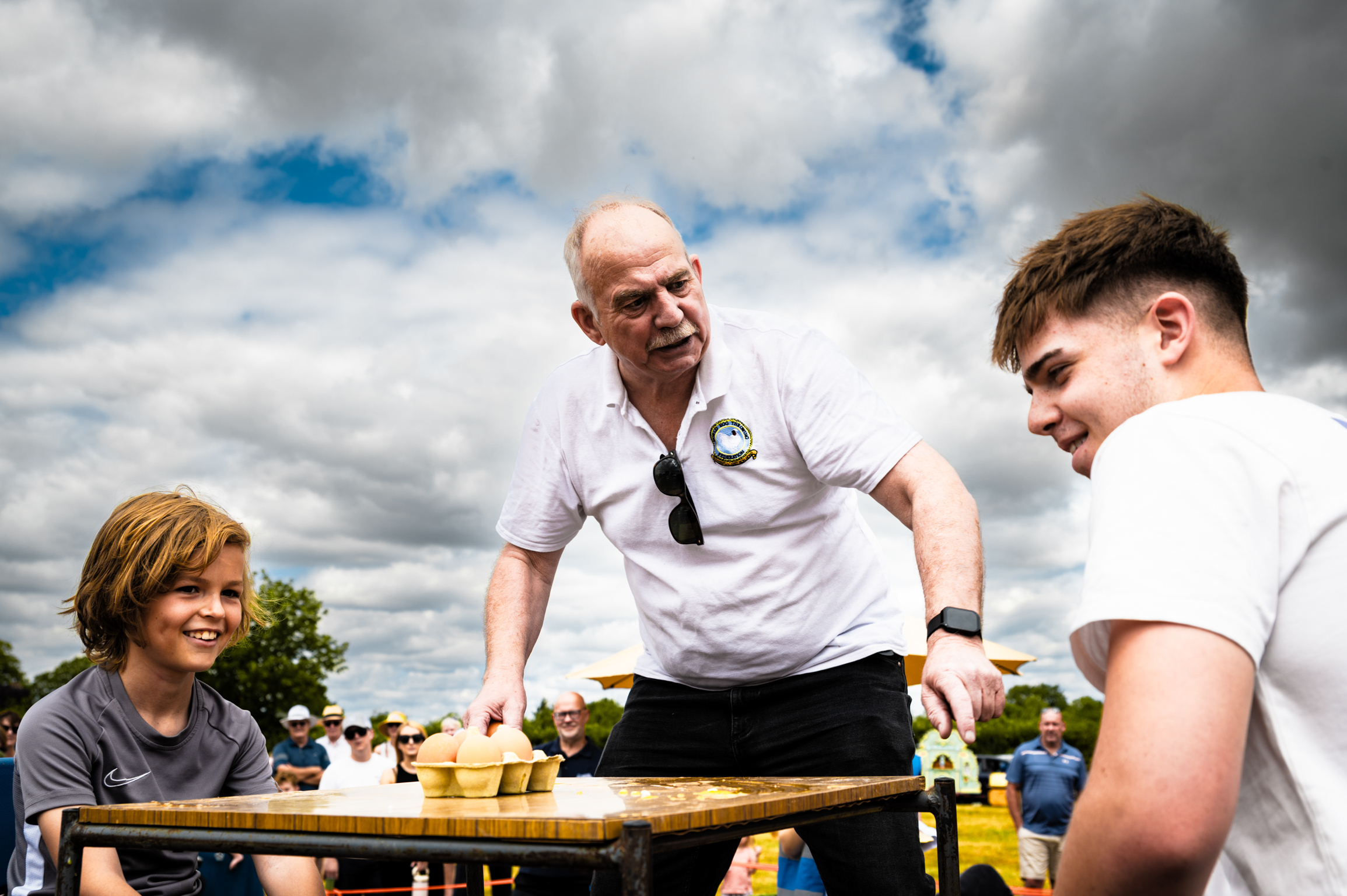 Bald man in white polo shirt stands at wooden table with two young people under cloudy blue sky at outdoor event.