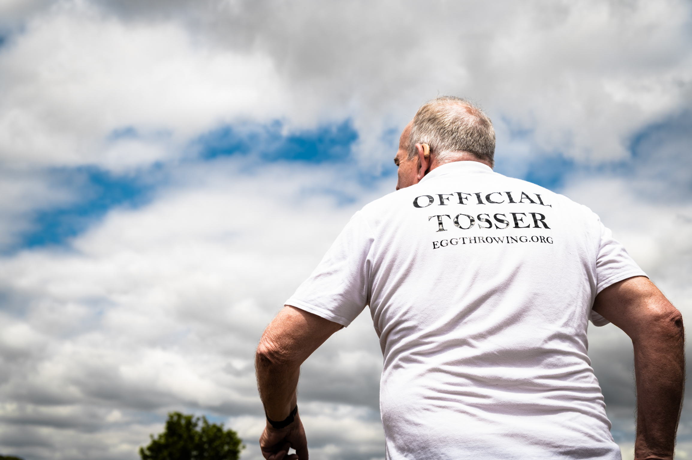 Man in white "Official Tosser" t-shirt viewed from behind against cloudy sky, leaning on railing with trees visible below.