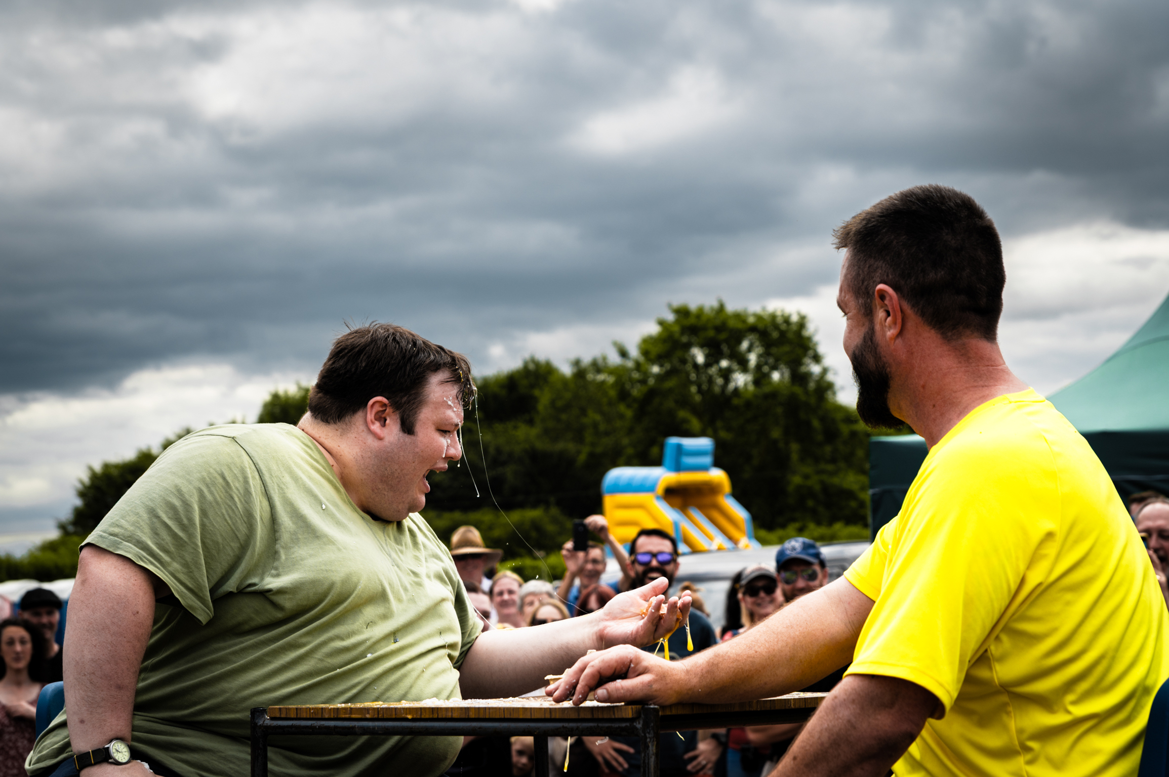 Two men arm wrestling at outdoor event, one in green shirt, one in yellow, with crowd and inflatable slide in background.