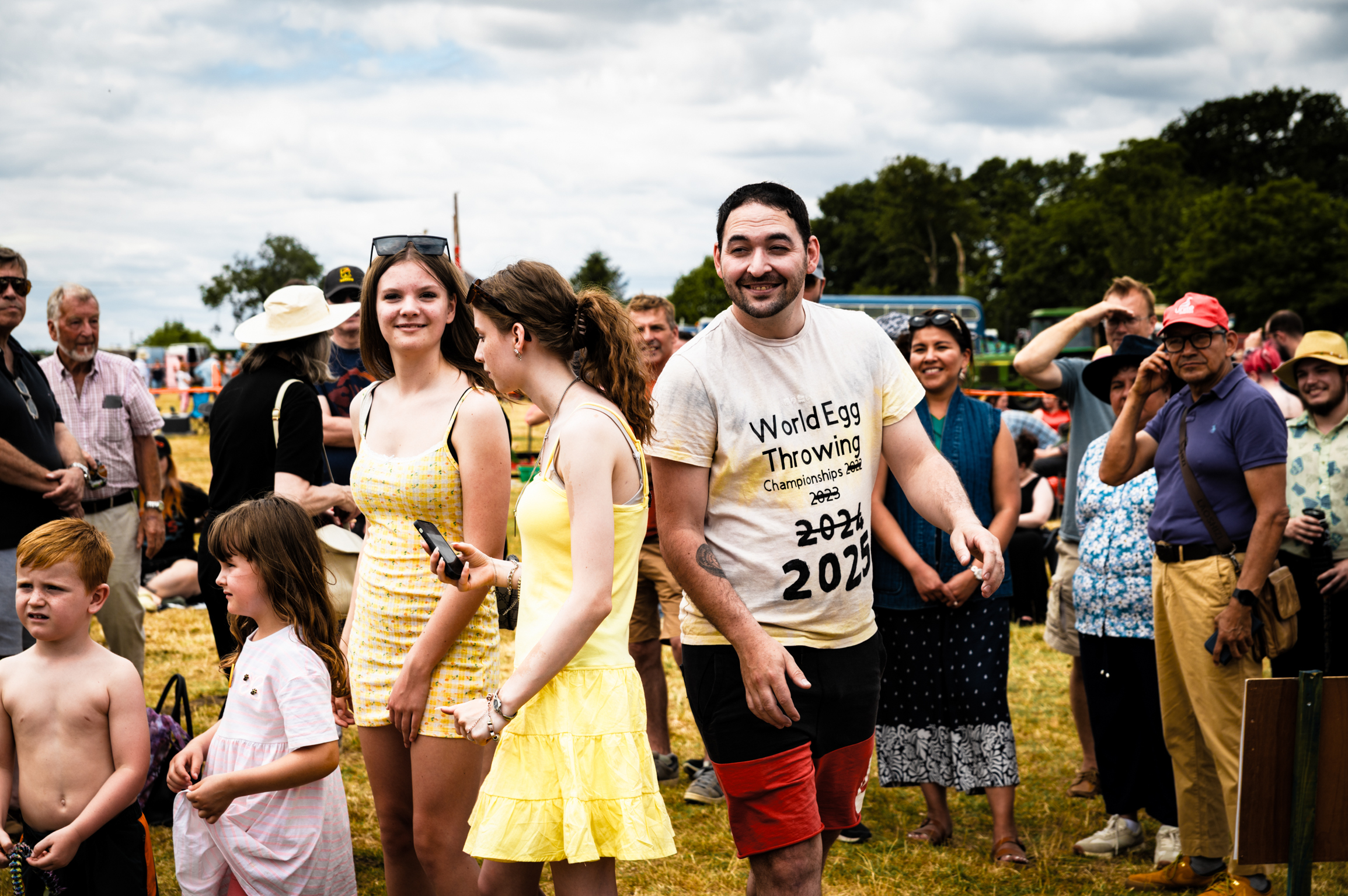 Group of people at outdoor event on grass field. Man in centre wearing white "World Egg Throwing" competition t-shirt, surrounded by spectators including children and adults in summer clothing.