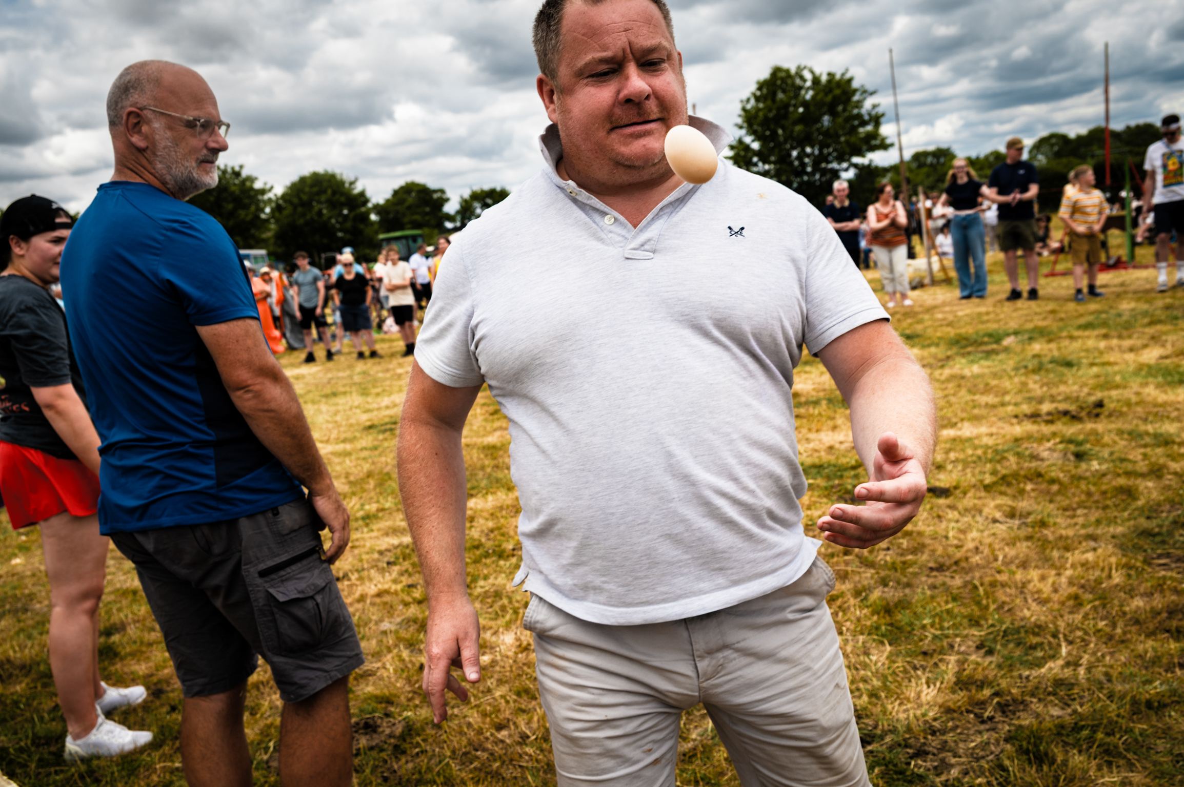 Two men at outdoor event on dry grass field. Man in foreground wears white polo shirt and beige trousers, man behind in blue shirt and dark shorts.