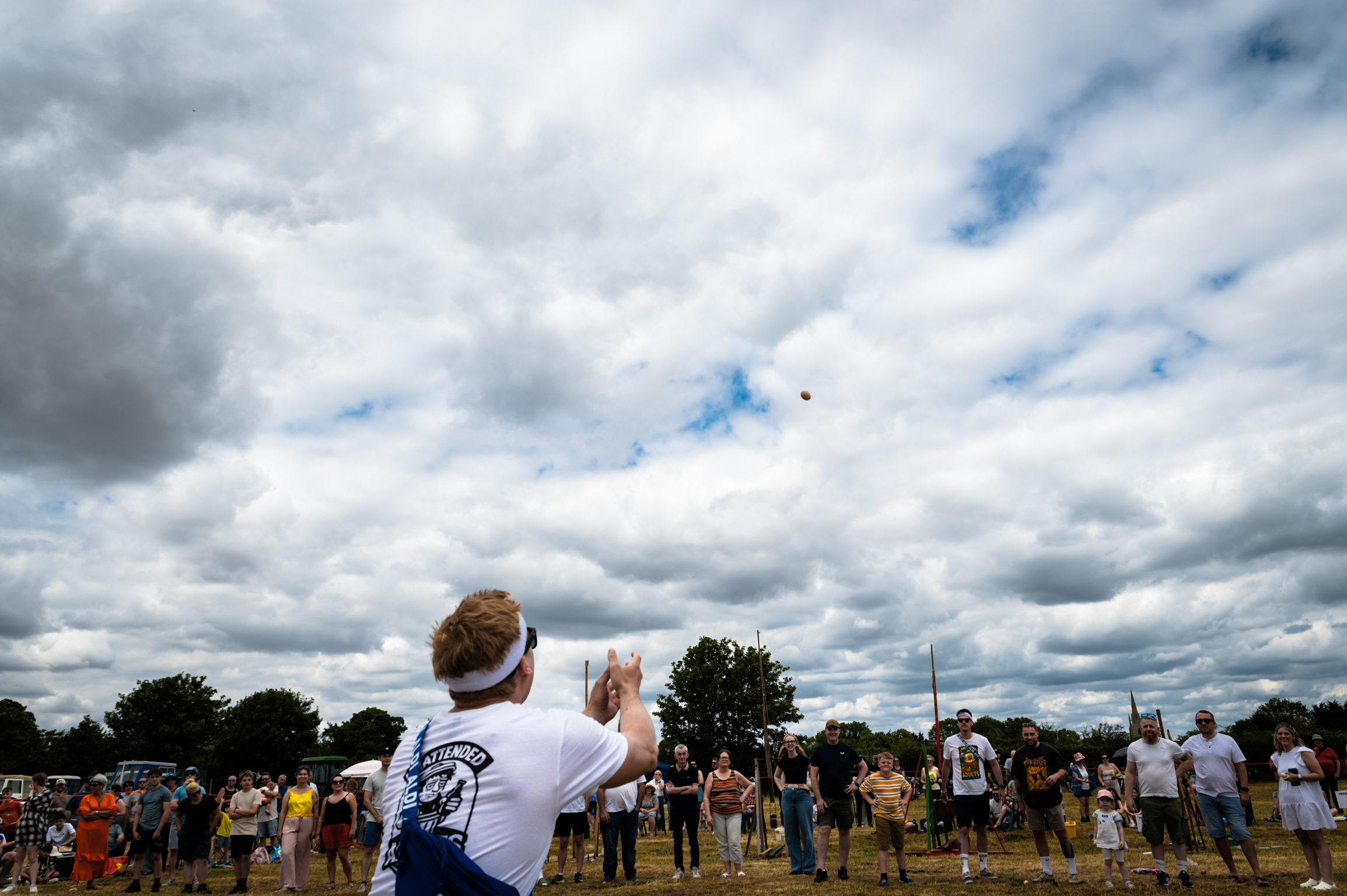 Man in white shirt with back to camera at outdoor event under dramatic cloudy sky with crowd of people scattered across field.