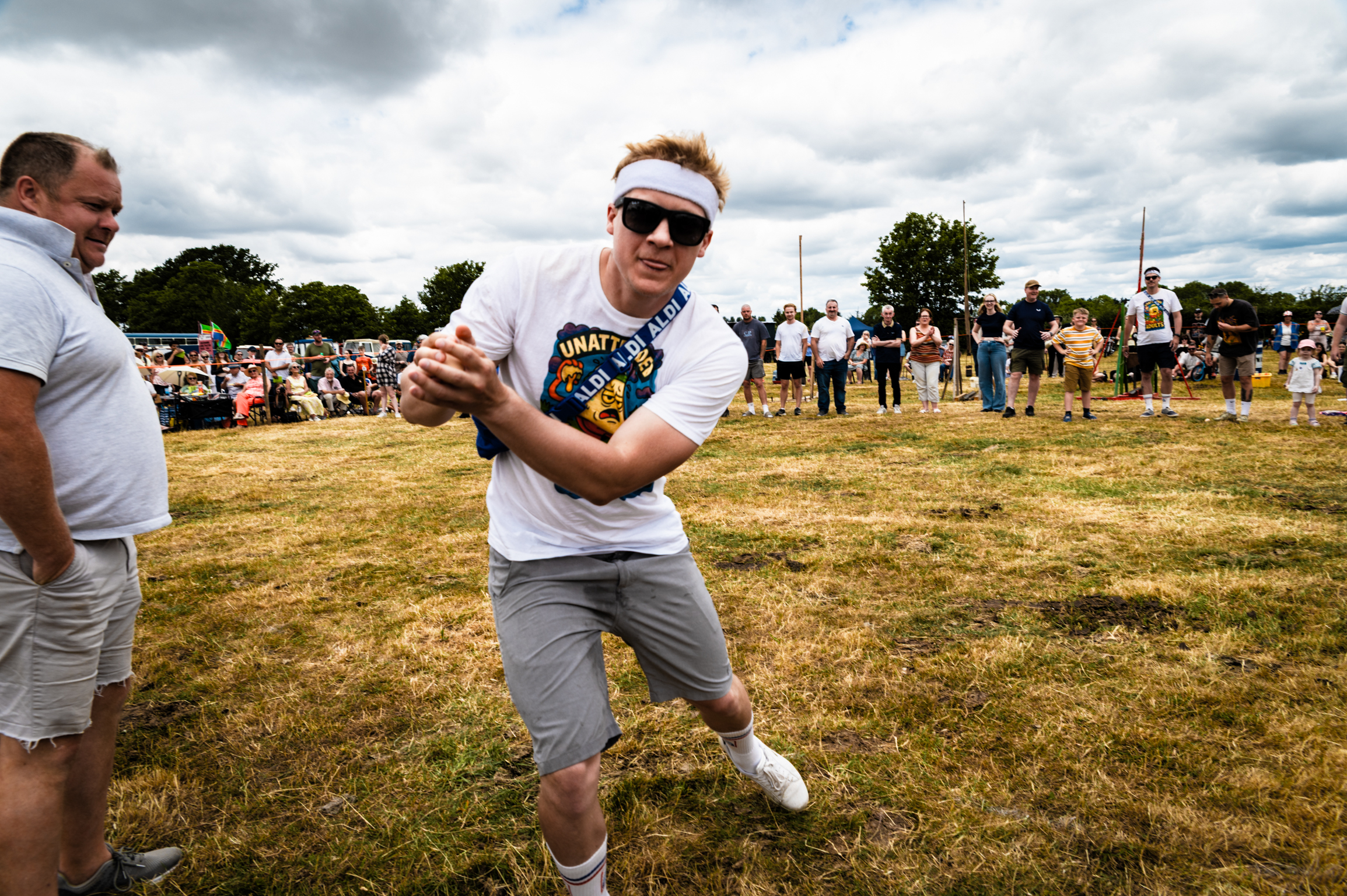 Man in white visor and t-shirt pulling rope during tug-of-war on grass field with crowd and cloudy sky in background.
