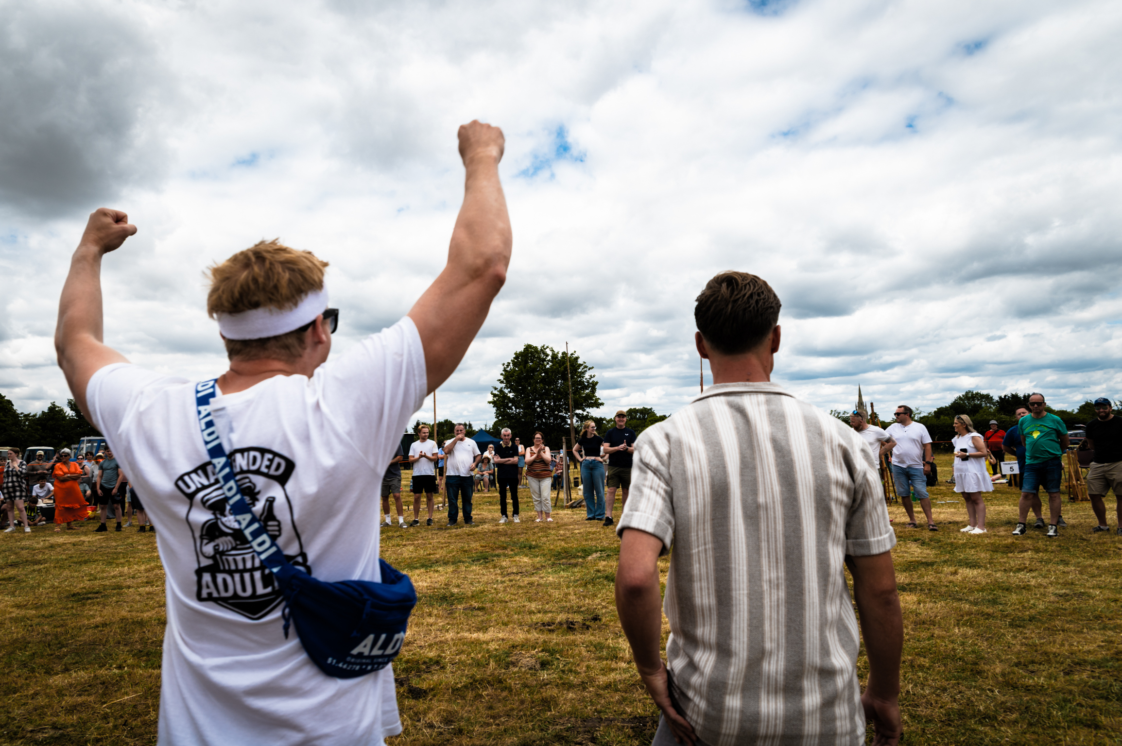 Two men with raised fists celebrating on grass field, crowd and cloudy sky in background at outdoor event.