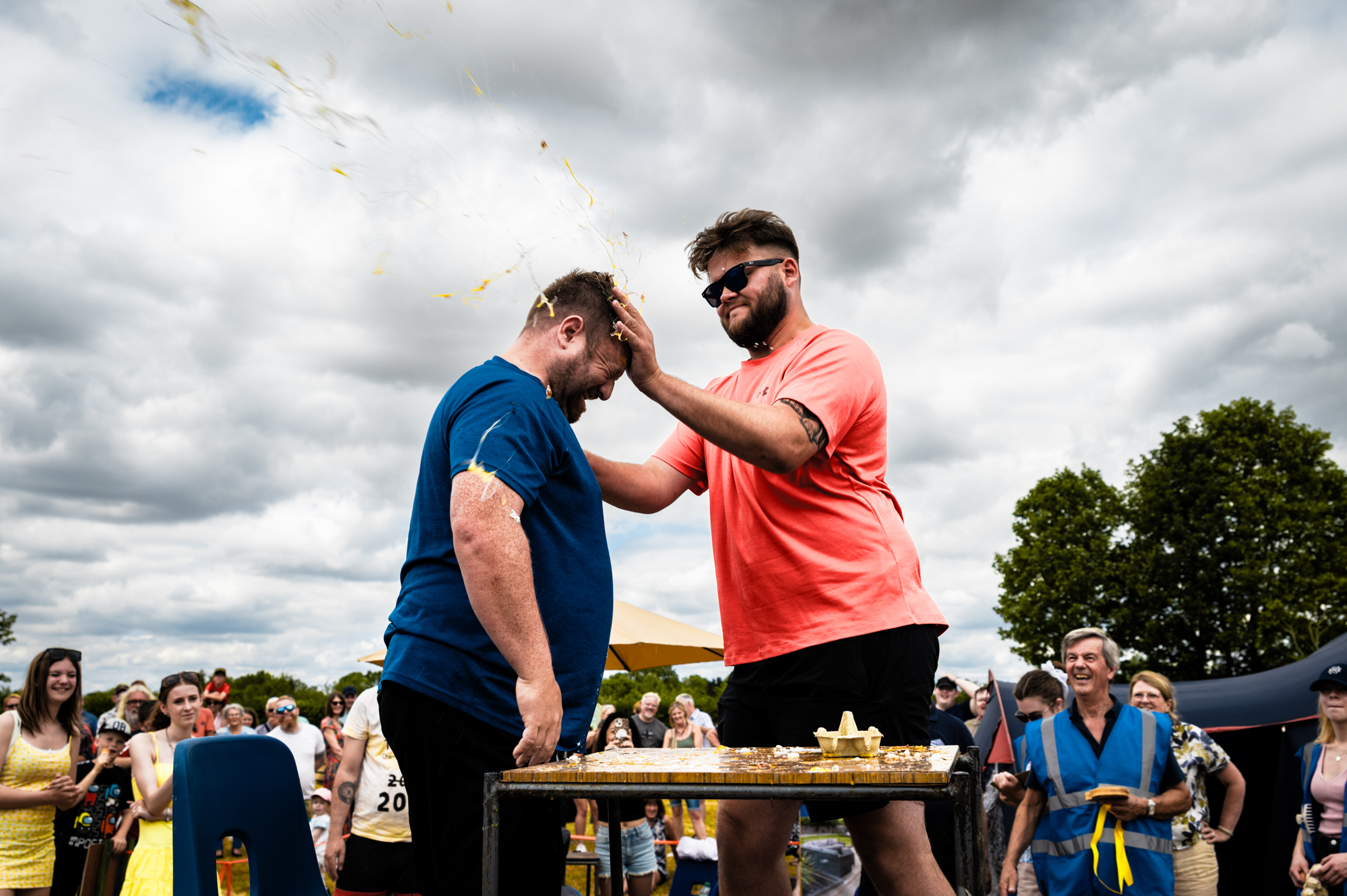 Man in orange shirt places medal around neck of man in blue shirt at outdoor event with crowd watching under cloudy sky.