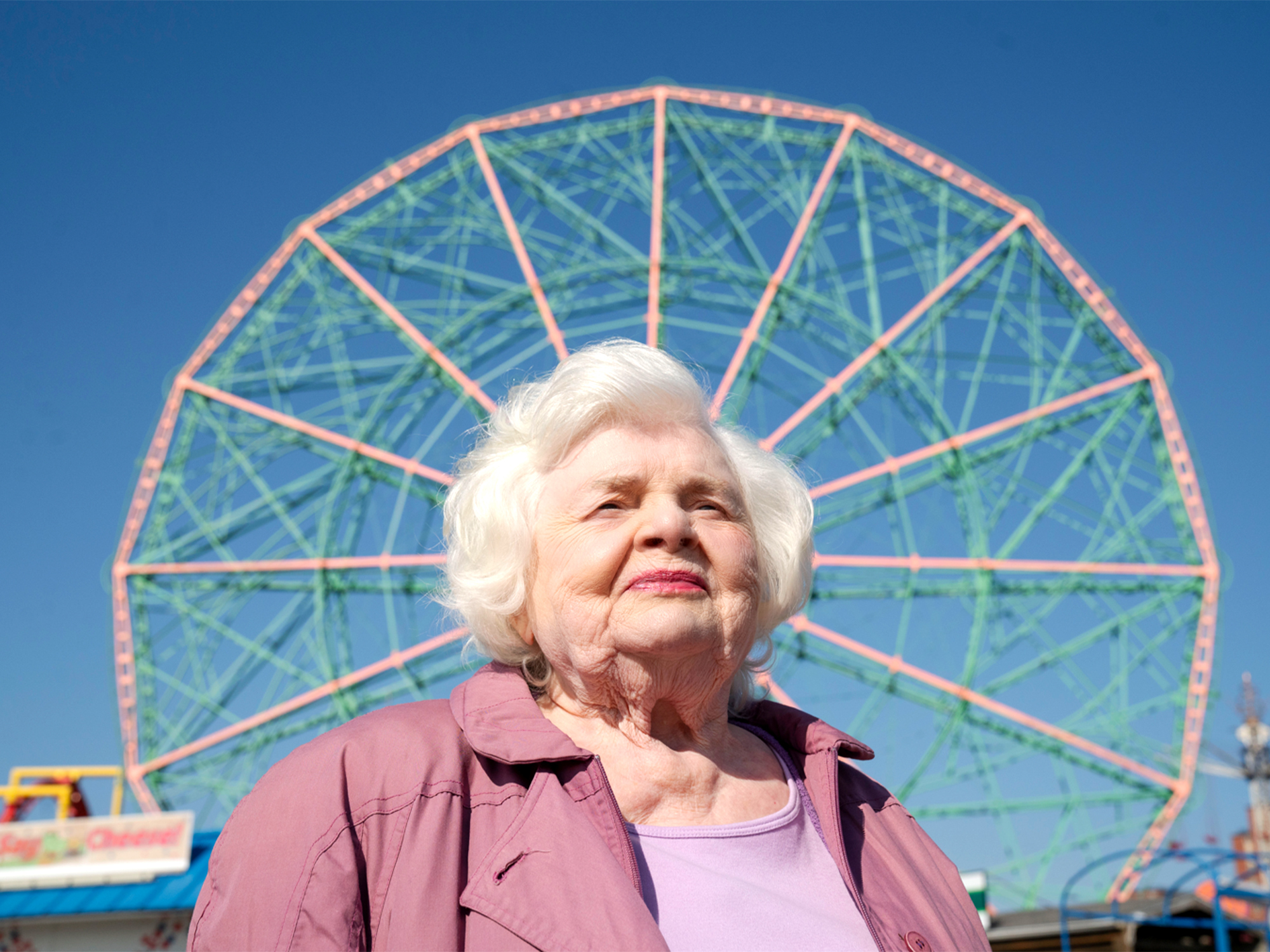 An elderly woman with white hair wearing a purple jacket, standing in front of a large colourful Ferris wheel.