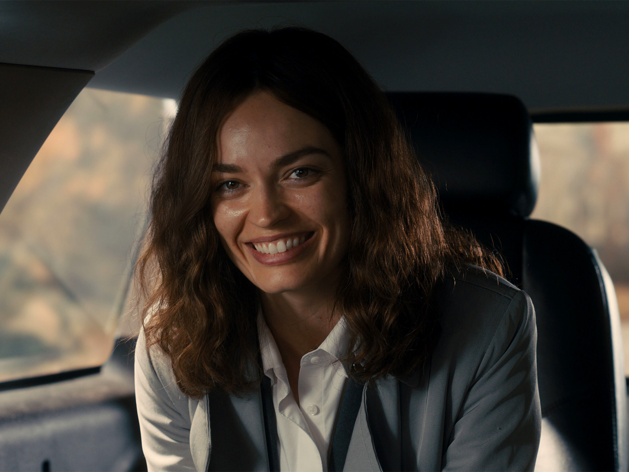 Woman with shoulder-length brown hair smiling in car interior, wearing white shirt and dark blazer, warm lighting through windows.