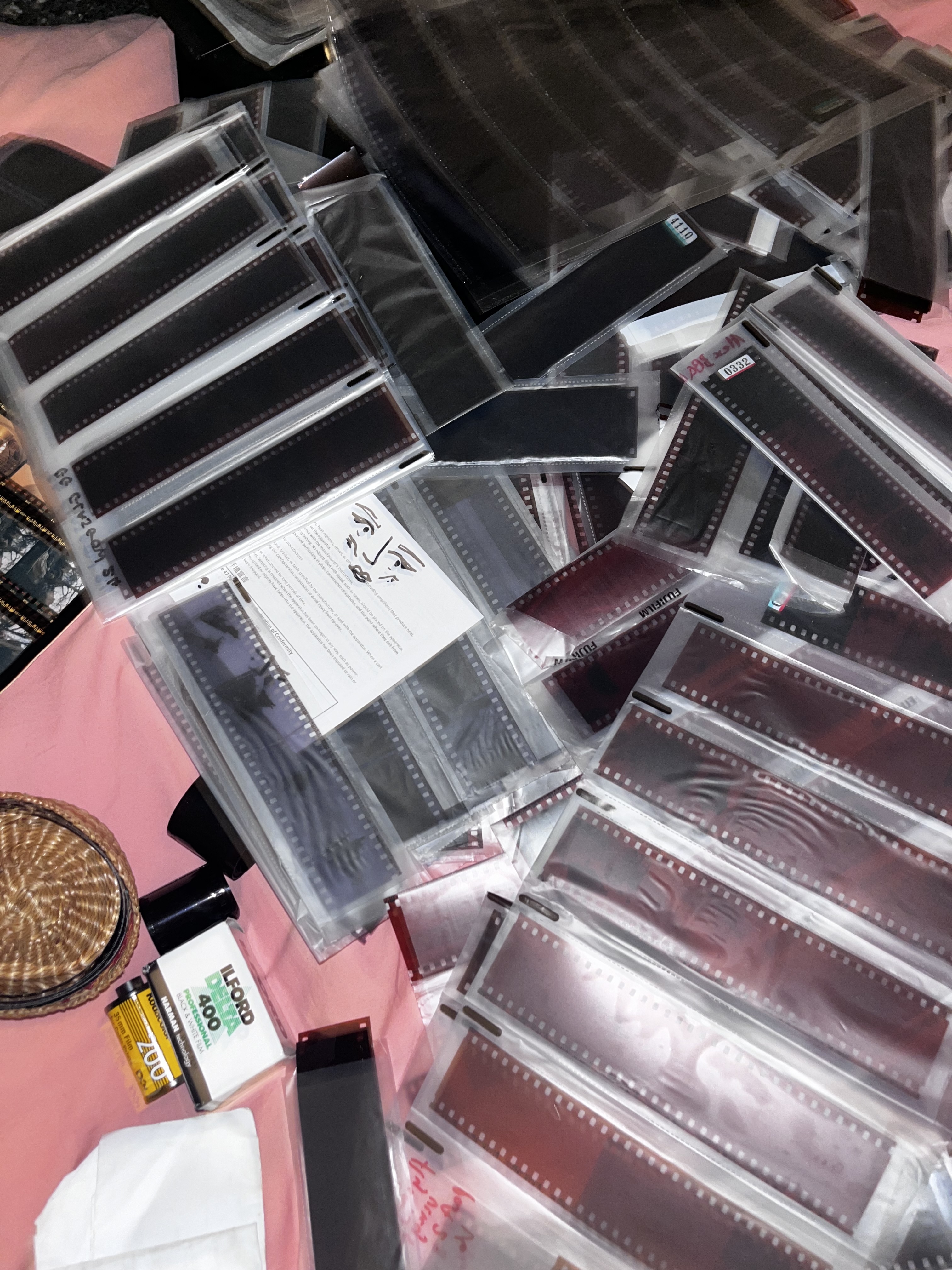 Collection of photographic film negatives in plastic sleeves scattered on pink surface with film canisters and contact sheets.