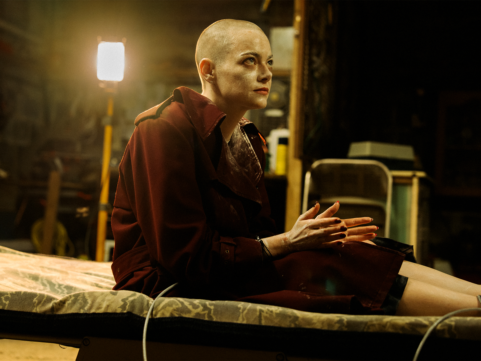 Bald person in dark red jacket sits on medical examination table with hands gesturing, bright light visible in background.