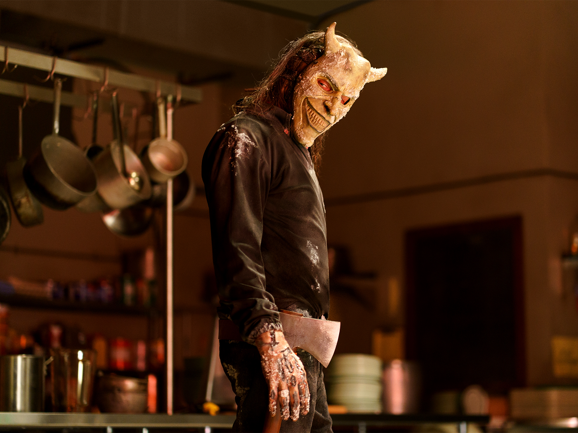 Person wearing horned demon mask and black clothing standing in kitchen with hanging pots and shelves in background.