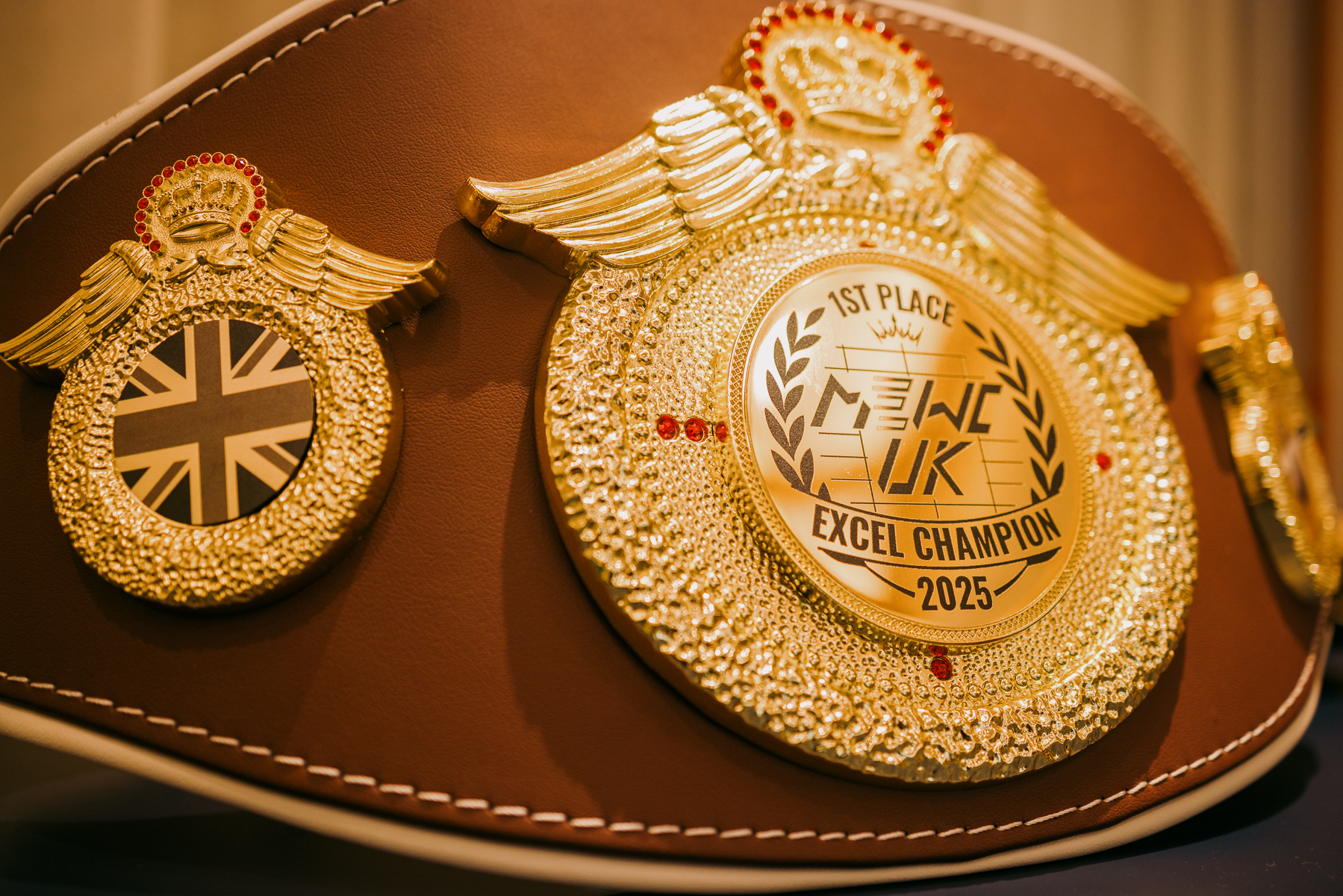 Golden championship belt with ornate circular medallions, winged decorations, Union Jack emblem, and "Excel Champion 2025" text on brown leather.